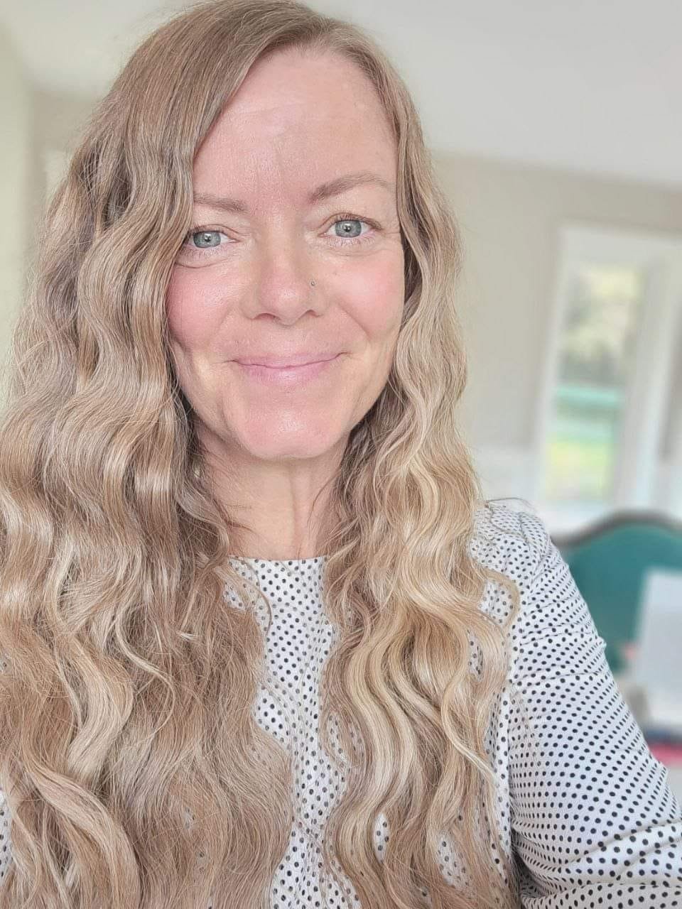 A woman with long wavy blonde hair and blue eyes smiling indoors.