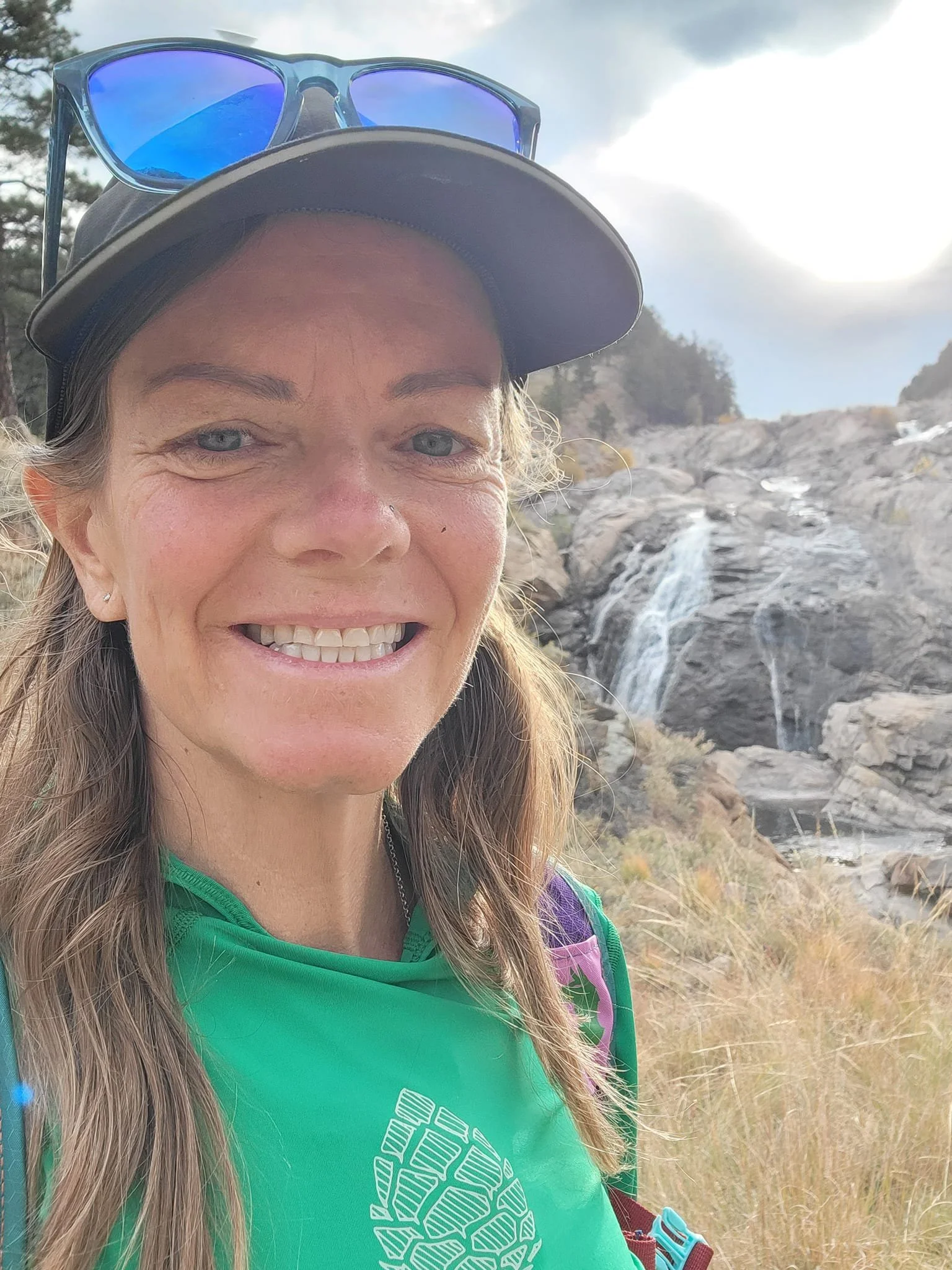 A smiling woman with long light brown hair, wearing a black cap with blue sunglasses on top, a green shirt with a white geometric design, and a purple backpack, standing outdoors near a rocky waterfall under a partly cloudy sky.
