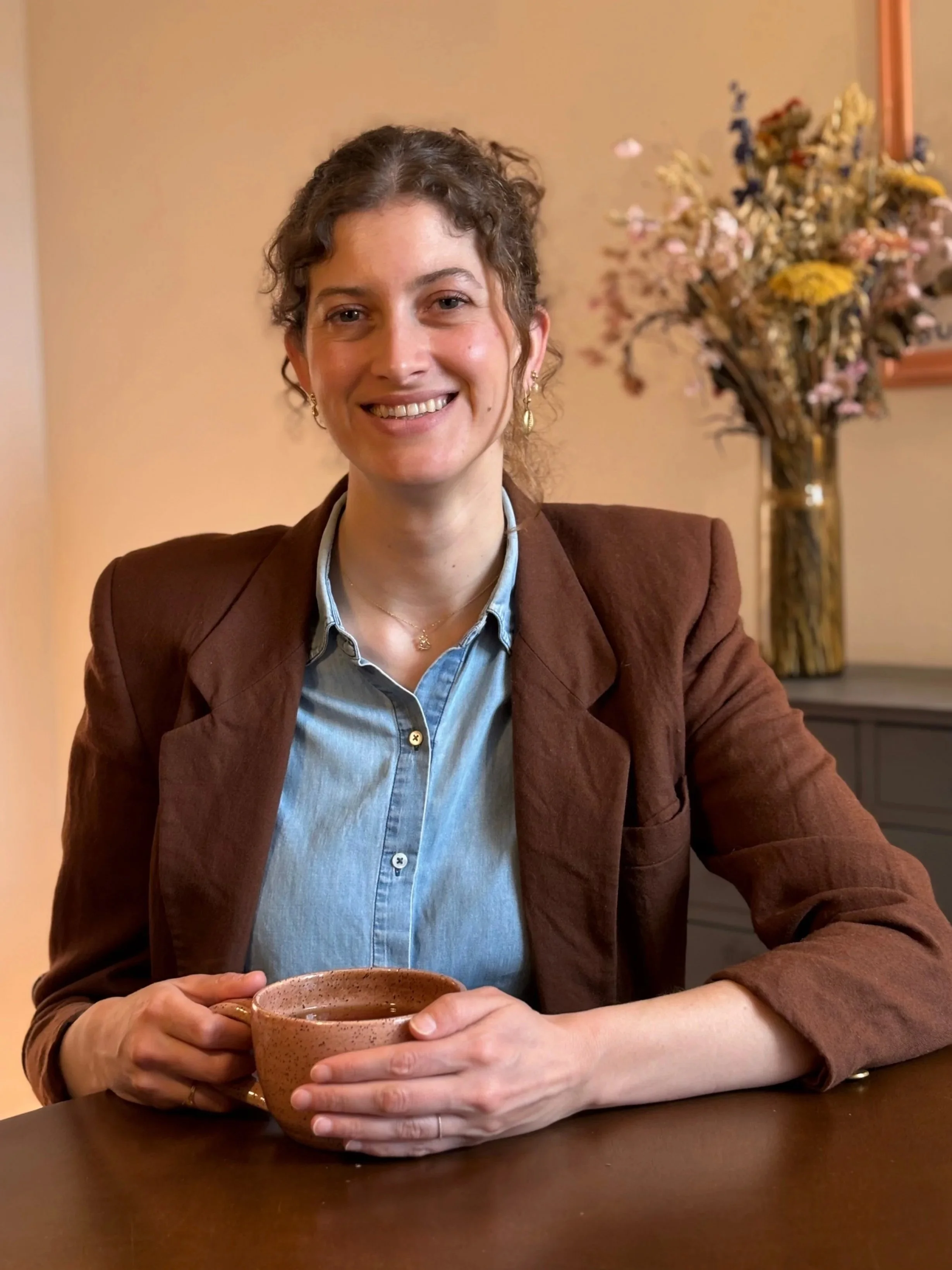 A woman with curly brown hair, wearing a brown blazer and light blue shirt, is sitting at a table holding a ceramic mug with both hands. There is a floral arrangement in a vase in the background.