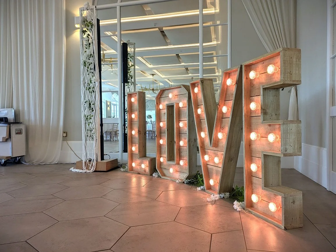Large illuminated marquee letters spelling 'LOVE' placed on a tiled floor in an elegant room with white walls, curtains, and a mirrored wall reflecting the space. Taken in the Orangery at Deer Park Country House