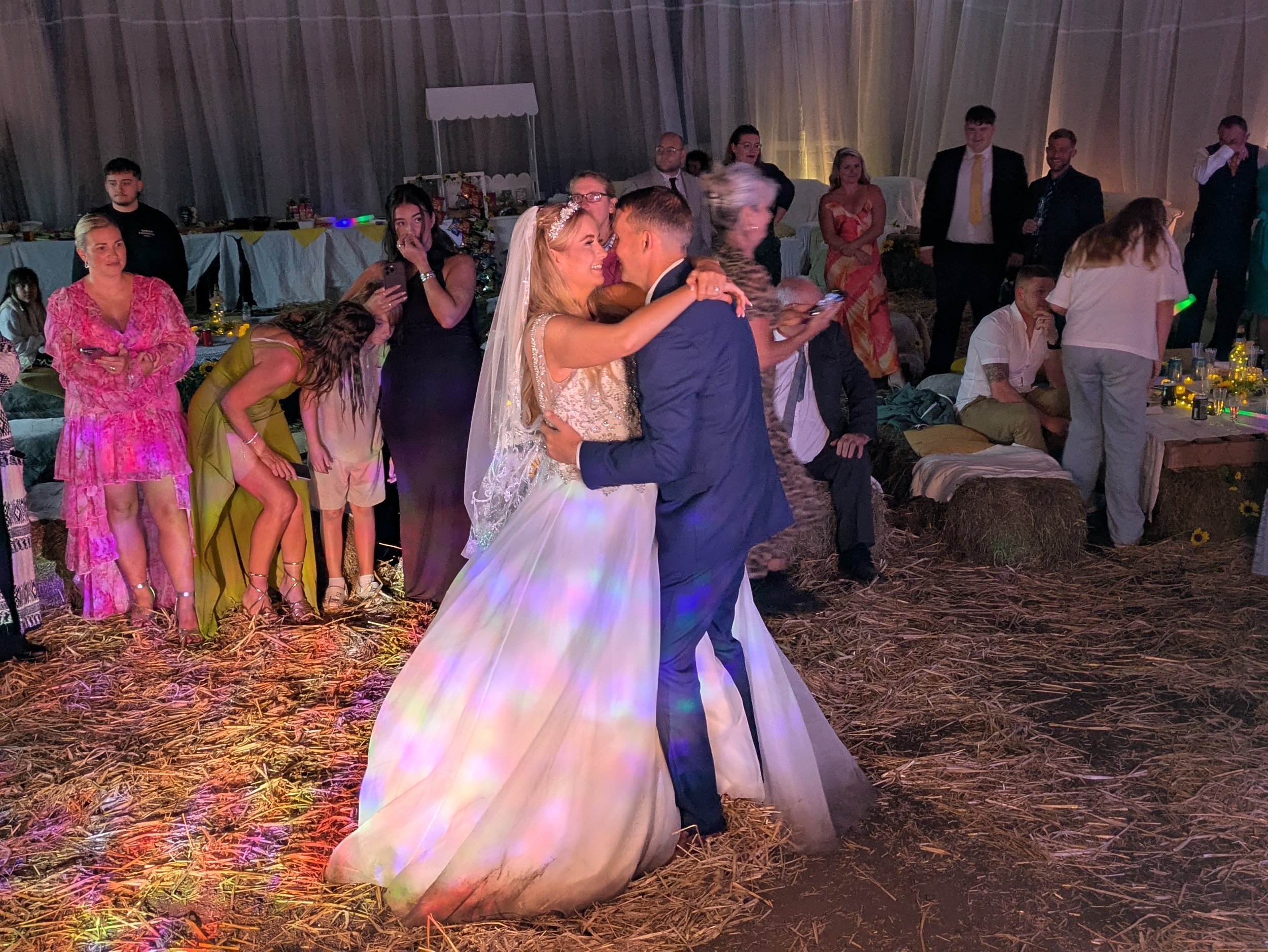 Beautiful couple having their first dance in a rustic barn, with family and friends all around sat on straw bales