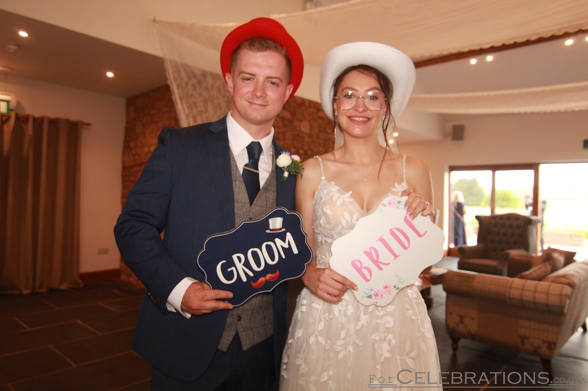 Bride and groom posing in a wedding PhotoPod, wearing fun hats and holding props during their reception at Quantock Lakes