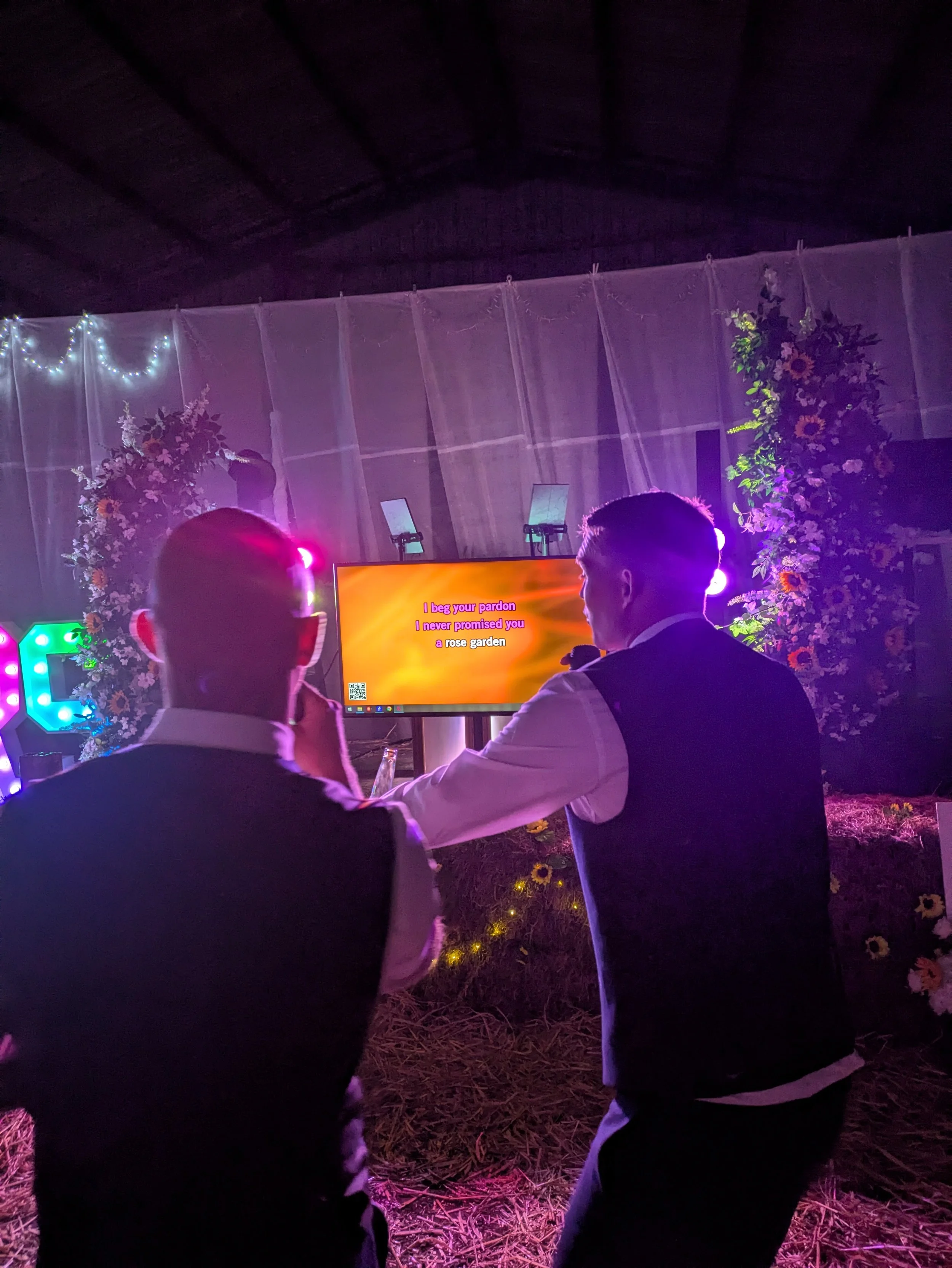 The groom and Best man singing karaoke in barn wedding venue with floral decorations, a large screen displaying song lyrics, and colorful lighting.