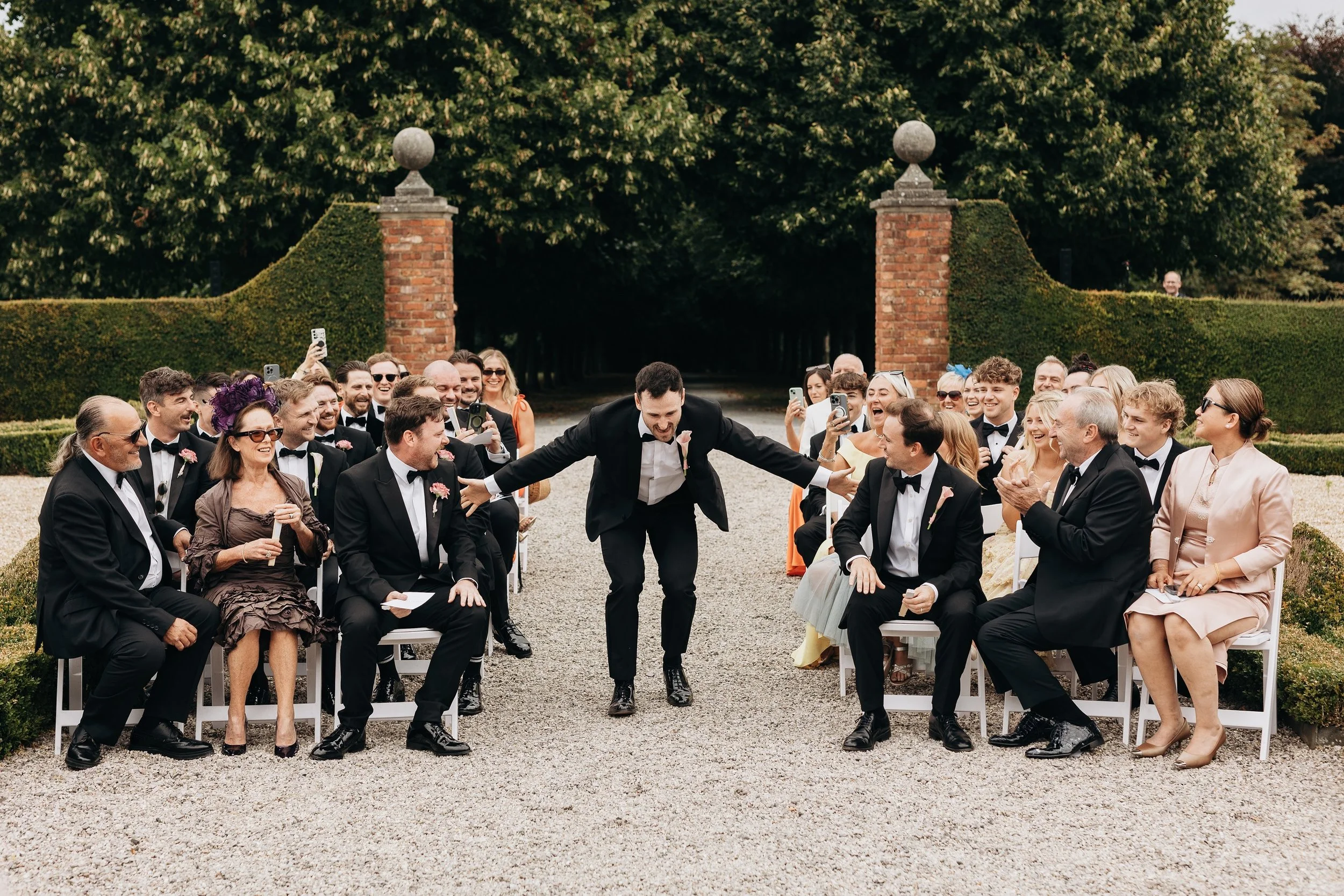 A wedding ceremony outdoors with guests seated on both sides of an aisle, celebrating as the groom bows with arms outstretched in front of the celebrant. The guests are clapping, smiling at Kingston Estate