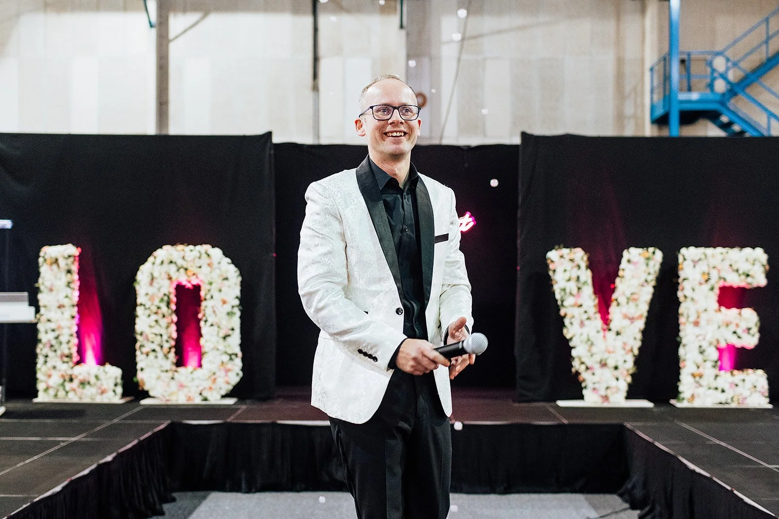 John William on stage, hosting the catwalk for Big South Wedding fair, dressed smartly with microphone and LOVE letters in the background