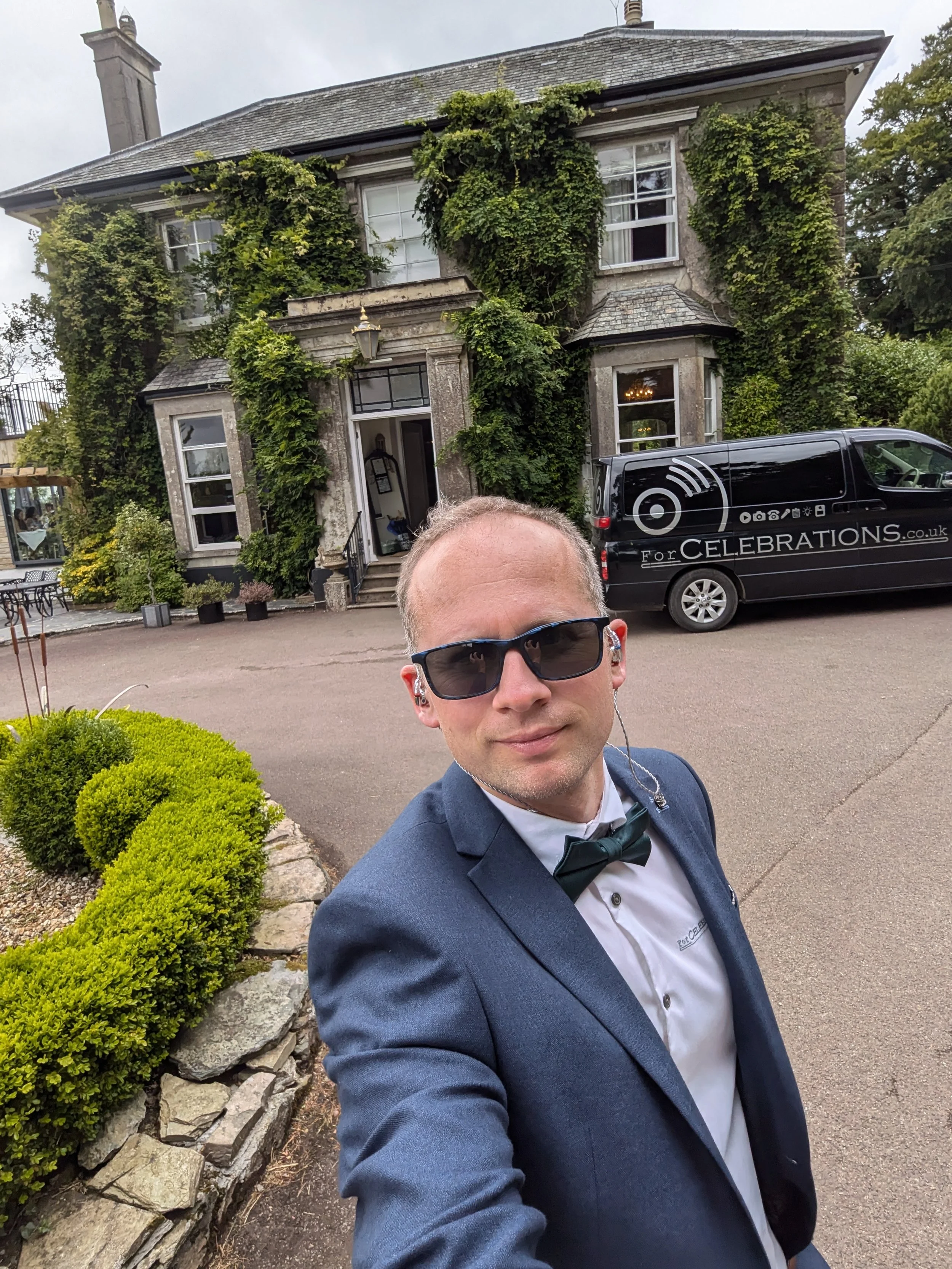 John William in a tuxedo and sunglasses taking a selfie in front of the Horn of Plenty, ivy-covered hotel, with a black van marked forcelebrations parked nearby.