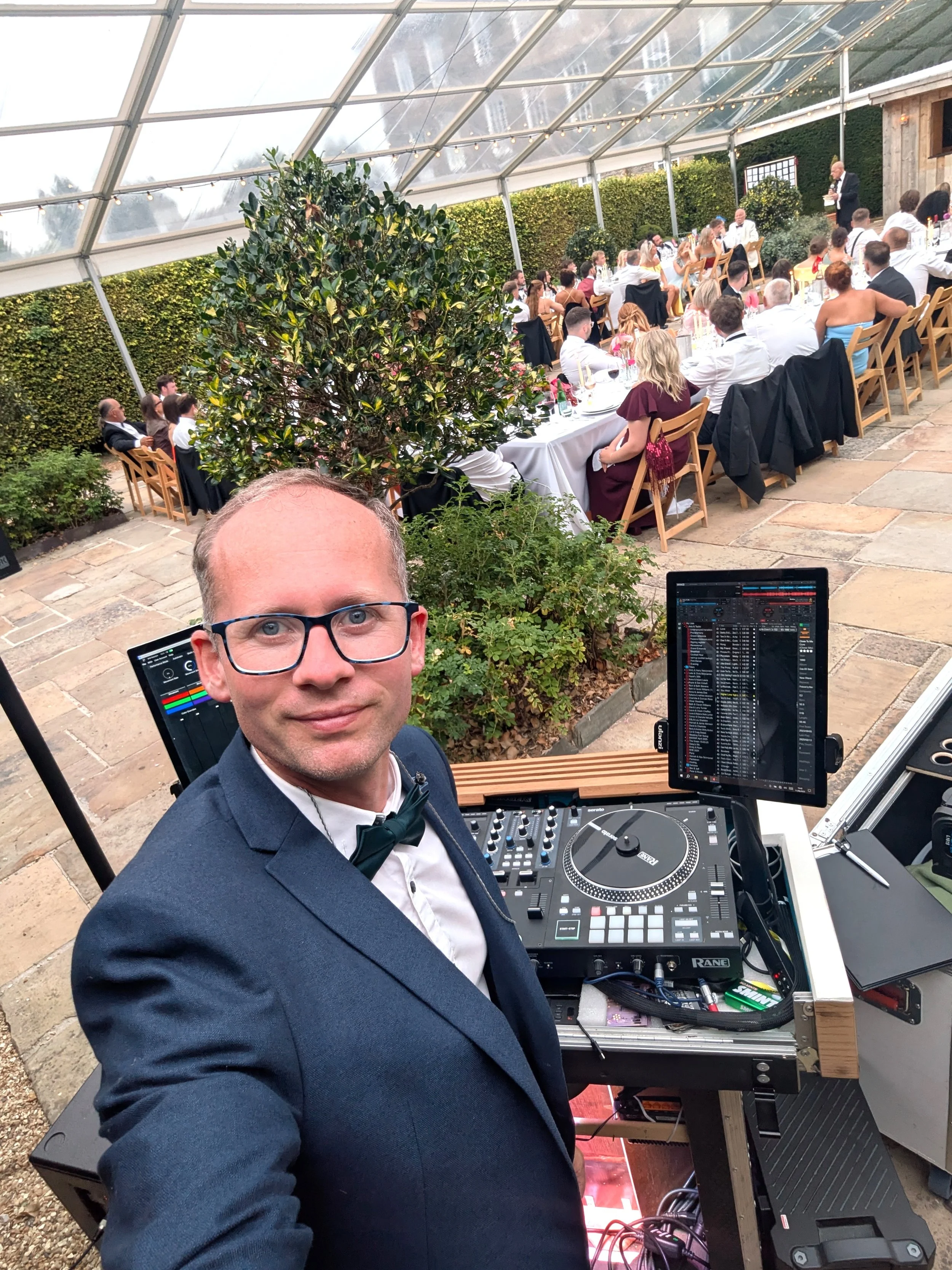John William, wedding host and DJ in a tuxedo and a bow tie taking a selfie at a wedding reception with guests seated at tables in the rose garden at Kingston Estate