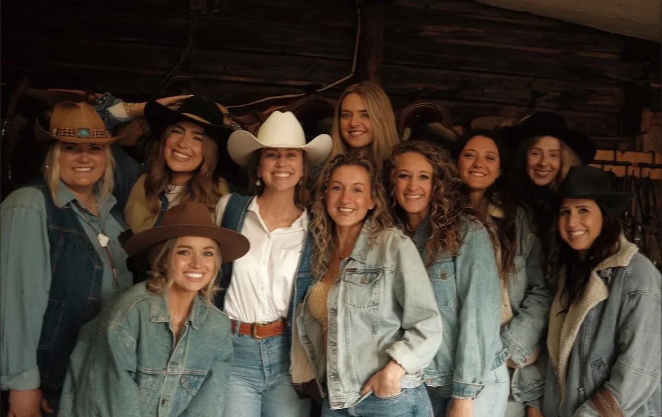 Group of women wearing cowboy hats and denim jackets inside a rustic wooden barn.