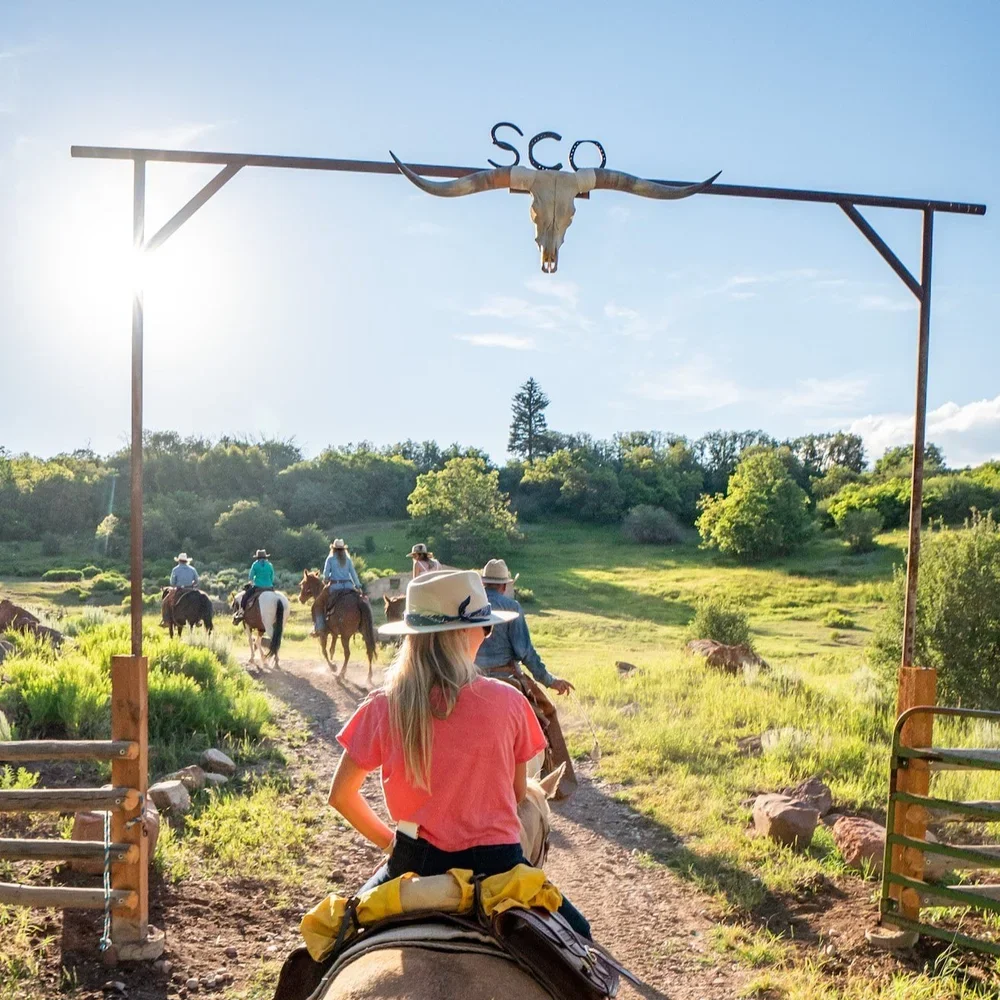 People horseback riding through a grassy trail on a sunny day, passing under an arch with a longhorn skull and the letters 'SCO' on top.