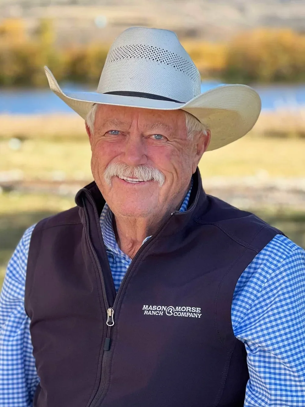 A senior man with a white cowboy hat, blue eyes, and a white mustache smiling outdoors, wearing a black Mason & Morse Ranch Company vest over a blue and white checkered shirt.