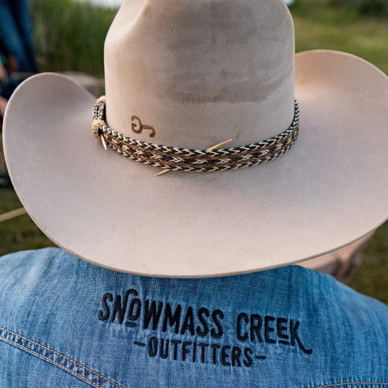 Back of a person's denim jacket with embroidered text 'Snowmass Creek Outfitters', wearing a beige cowboy hat with a braided band and a small decorative pin, outdoors.