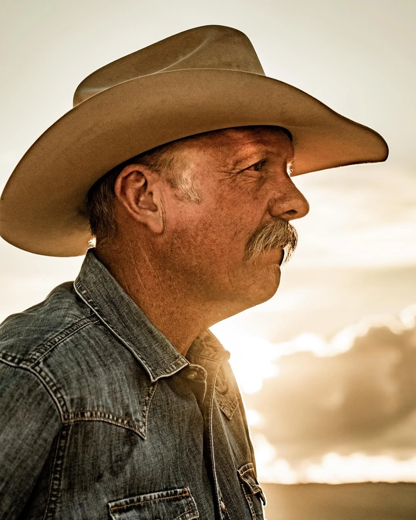 Profile of a man wearing a cowboy hat, denim shirt, at sunset or sunrise outside.