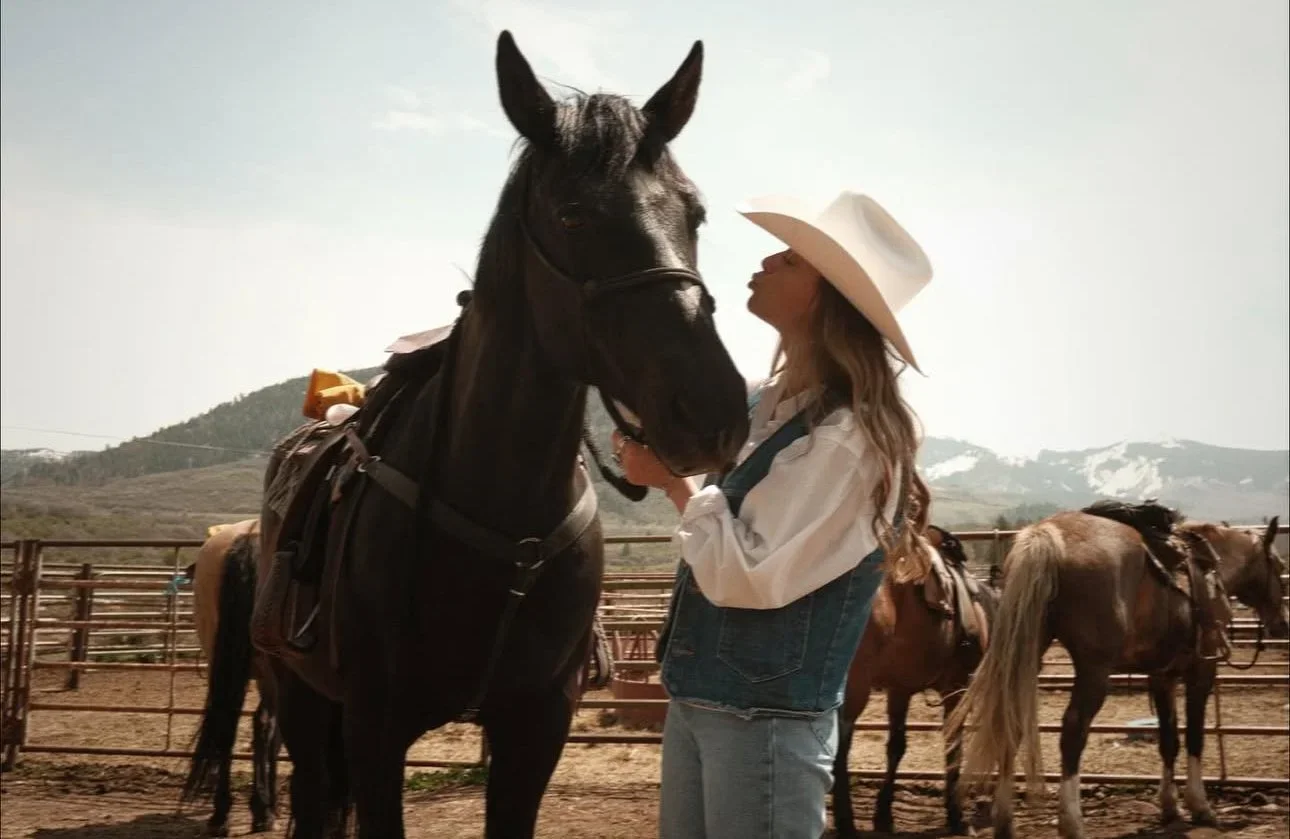 Woman with long hair wearing a white hat and denim vest touching a black horse in a riding arena on a sunny day.