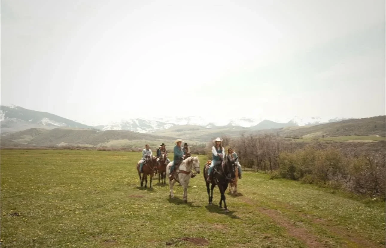 Group of five people riding horses in a grassy field with mountains and snow in the background.