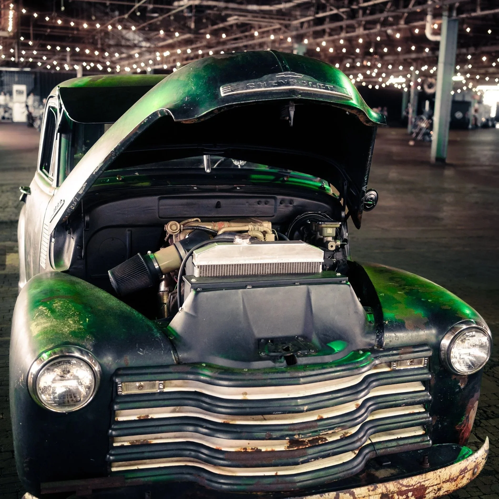 Front view of a vintage Chevrolet car with its hood open, showcasing the engine. The car appears weathered with signs of rust and chipped paint, and is parked in a large indoor space decorated with string lights.