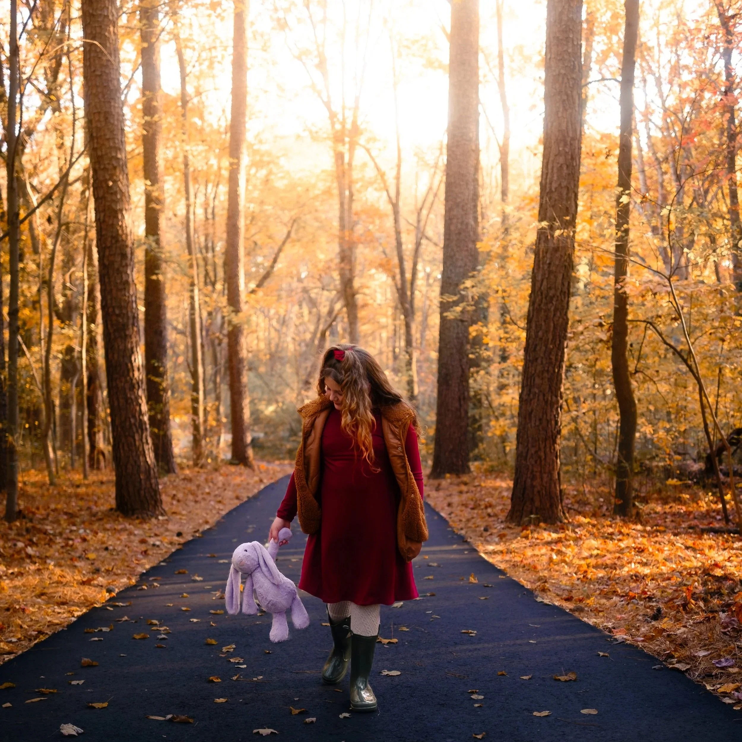 A young girl with curly hair walking on a paved trail through a forest with orange and yellow autumn leaves. She is wearing a red dress, a brown vest, grey leggings, and black boots, and is holding a purple stuffed bunny.