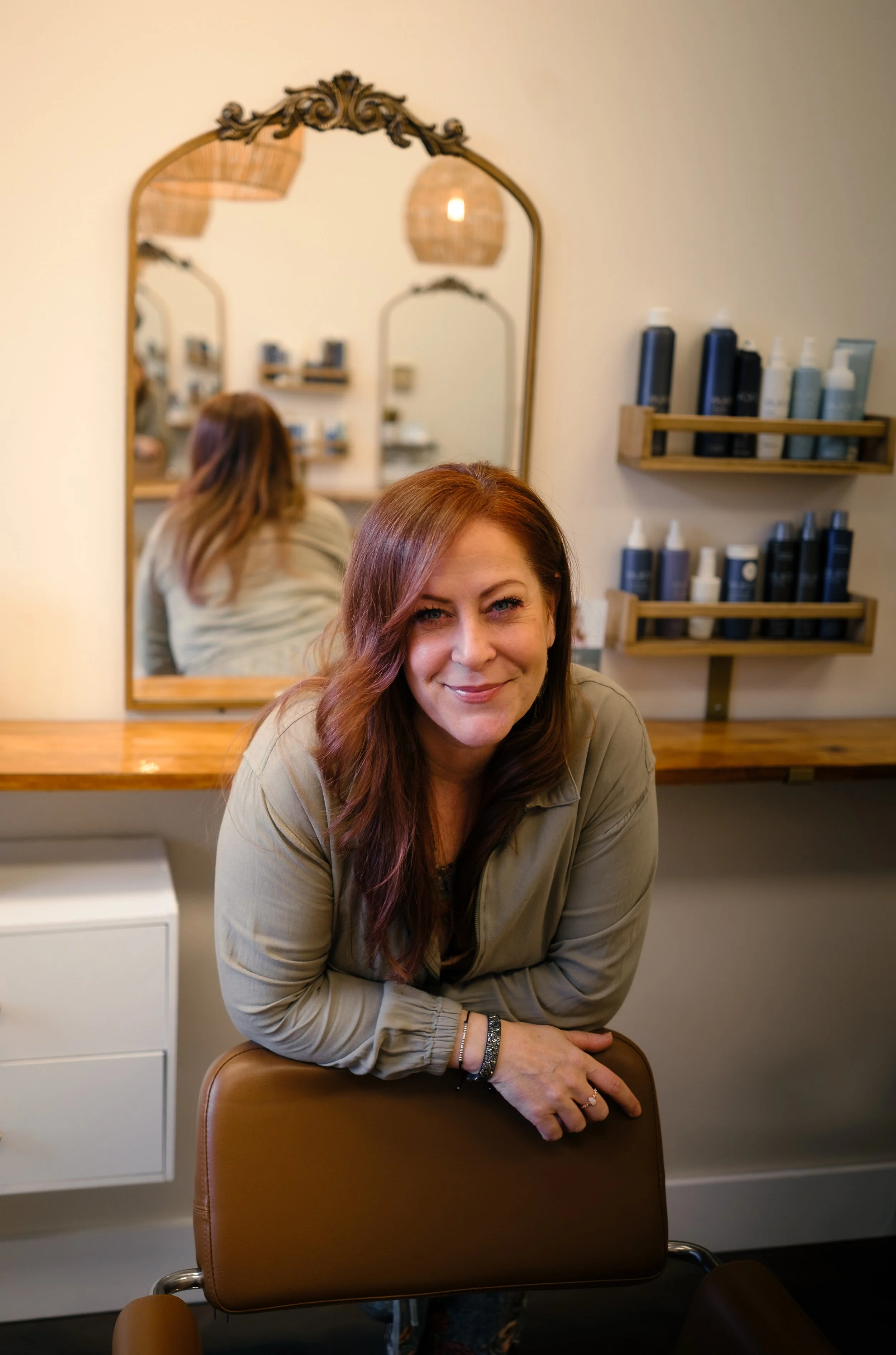A woman with long red hair sitting in a salon chair, smiling, with a mirror and shelves of hair products in the background.