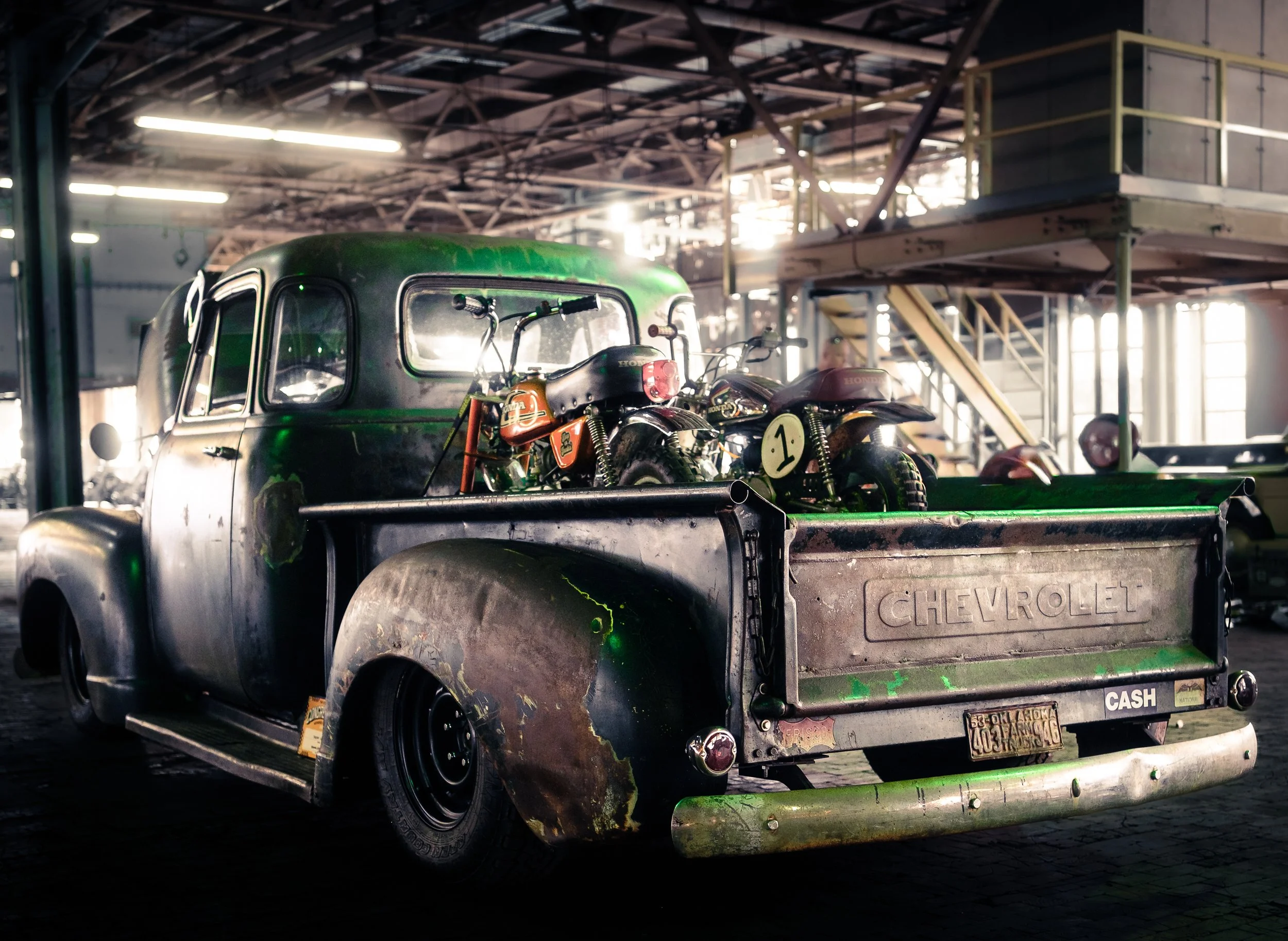 An old Chevrolet pickup truck with rusty and weathered body, containing two vintage motorcycles in the truck bed, inside a spacious industrial-style building with high ceilings and metal staircase.