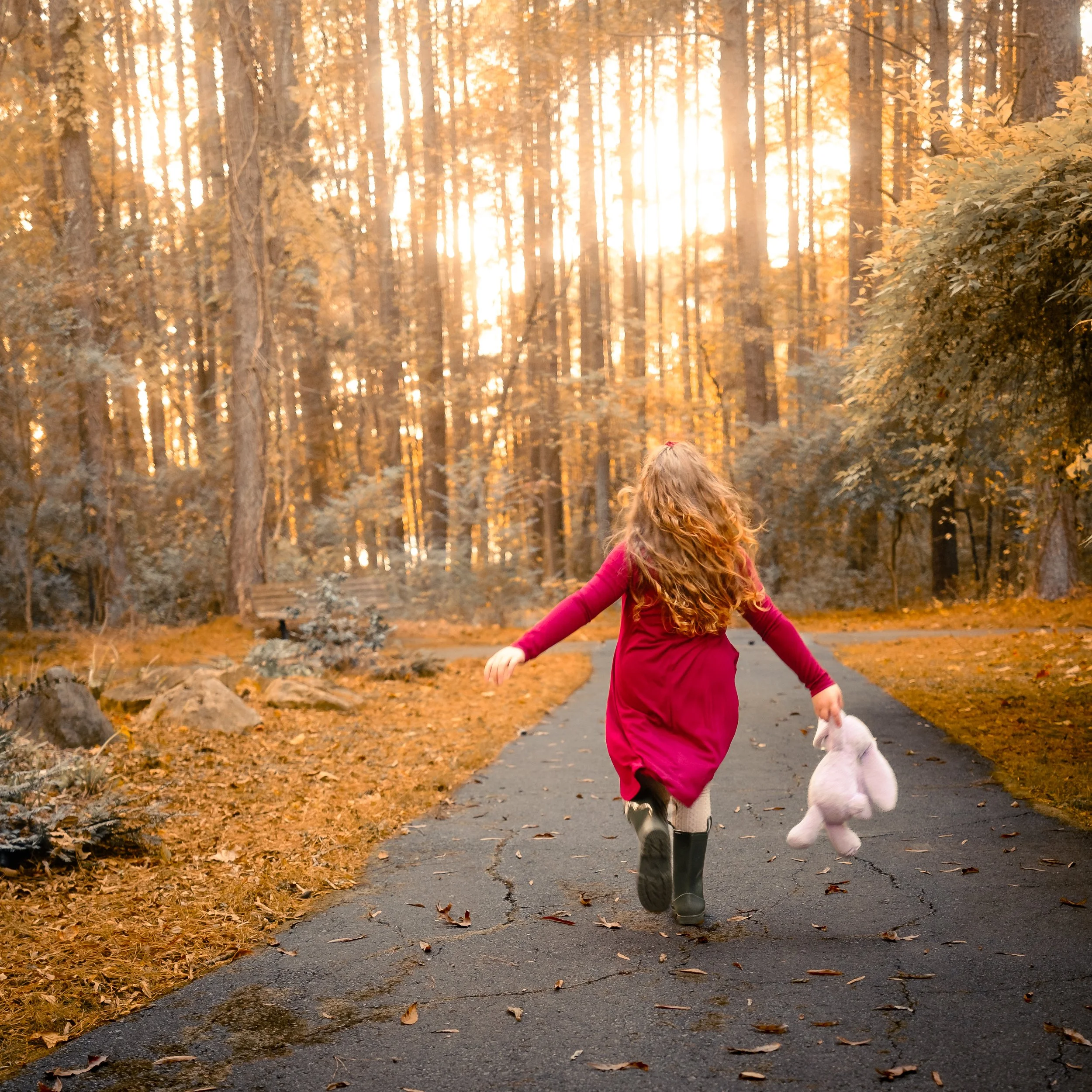 A girl in a red dress walking along a paved path in a forest during autumn, holding a white stuffed animal.