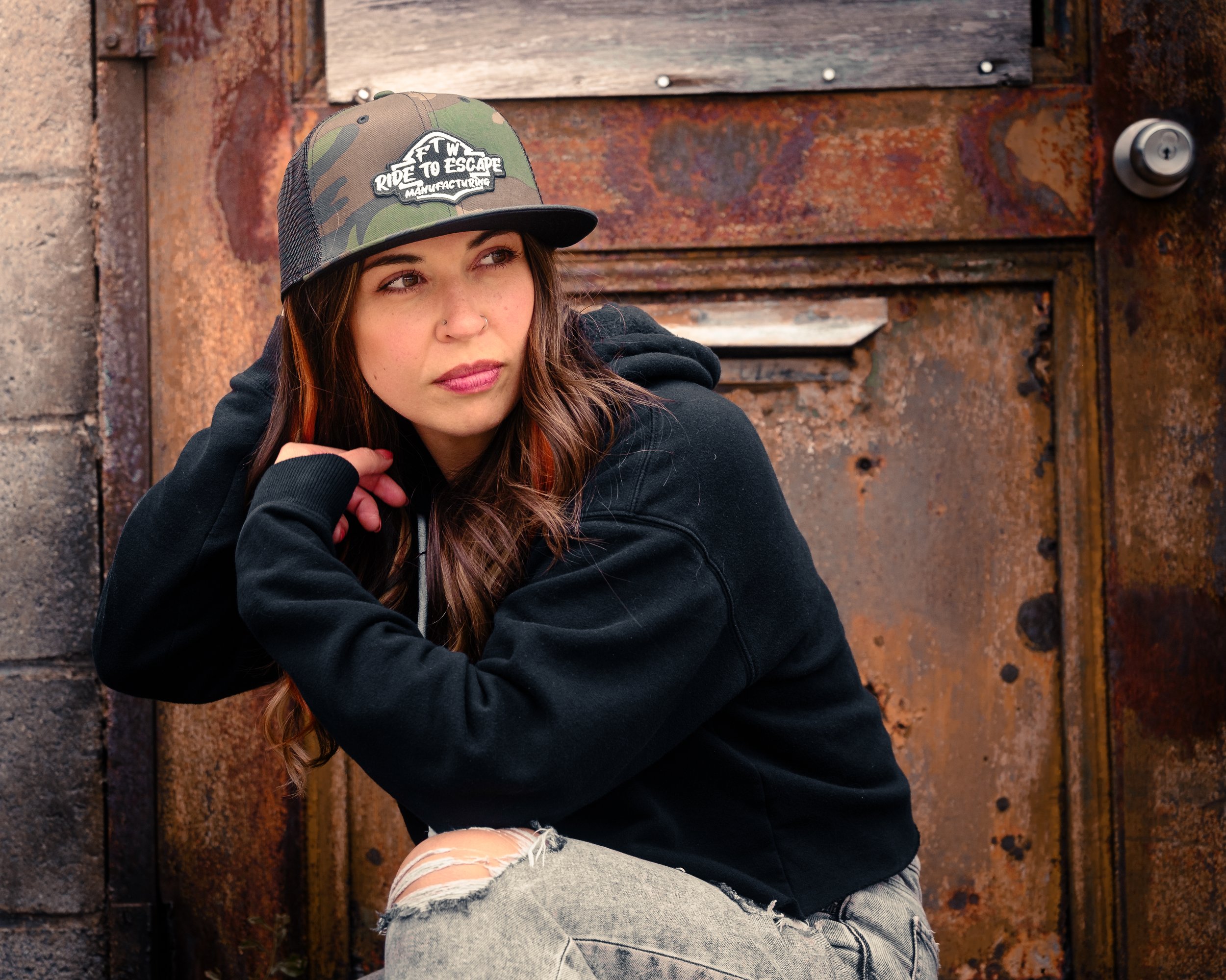 A young woman with long brown hair, wearing a black hoodie and a camo baseball cap with the words 'Ride to Escape Manufacturing' on it. She is crouching next to a rusty metal door with a lock, leaning on her left hand and looking to the right.