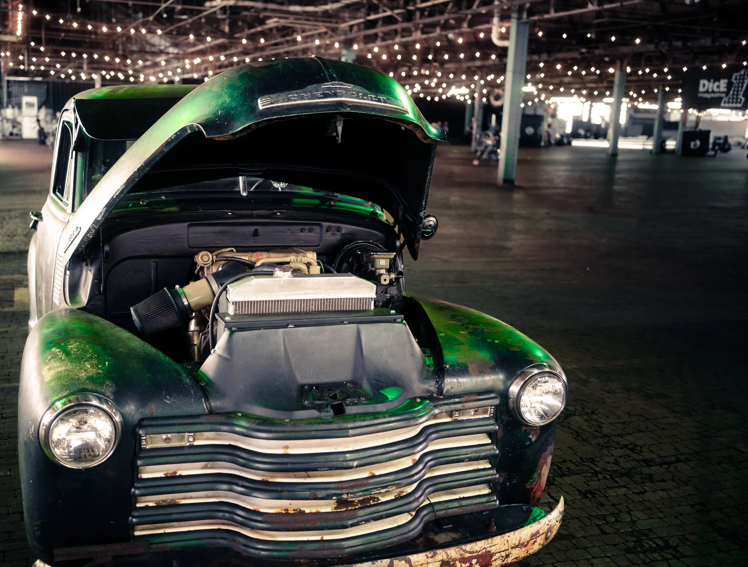 Vintage Chevrolet car with its hood open, showing the engine, parked inside a dimly lit, large indoor space with string lights hanging from the ceiling.