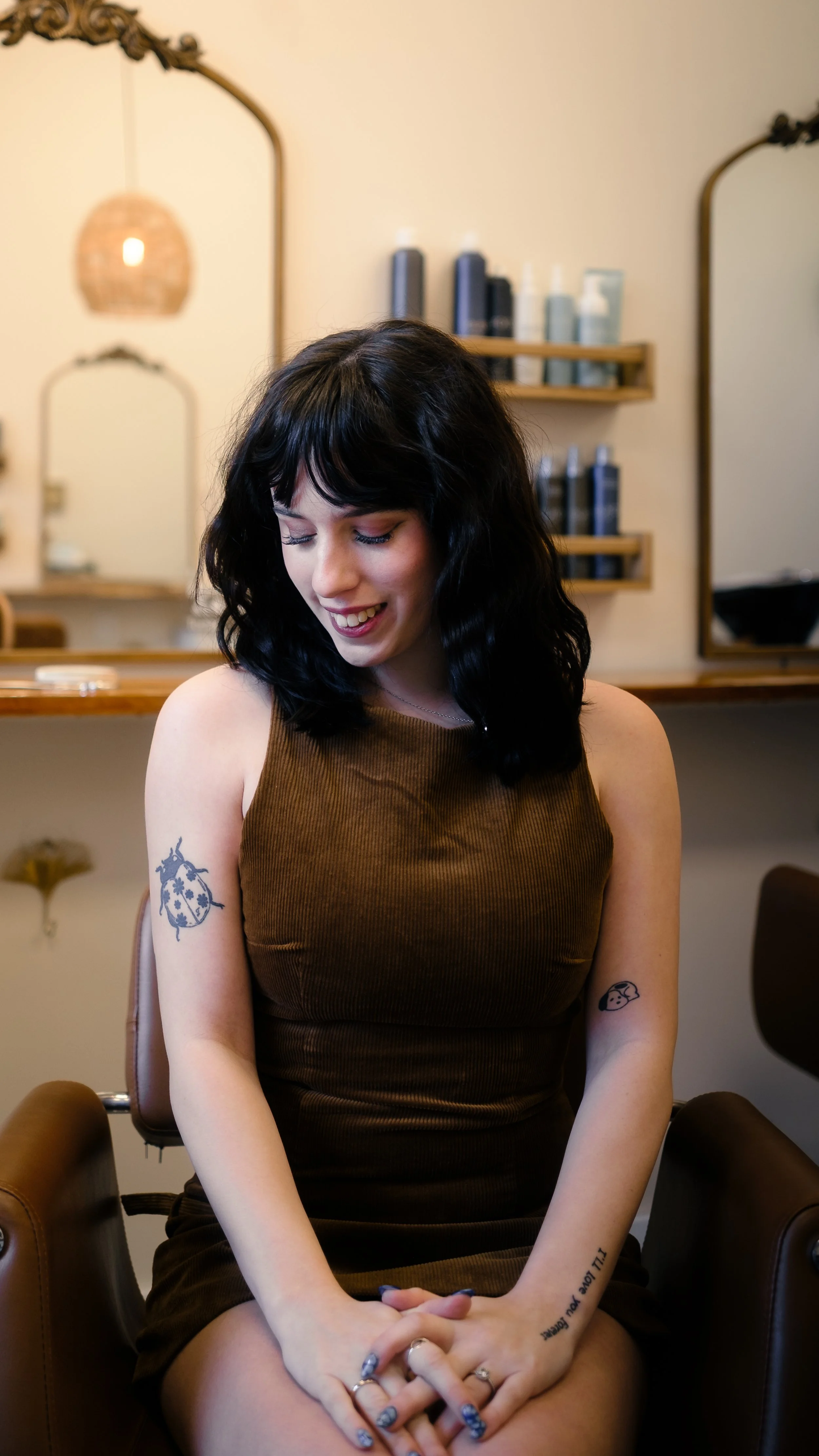 A young woman with black wavy hair, tattoos on her arms, and wearing a brown sleeveless dress, sitting in a salon chair and smiling with her eyes closed.