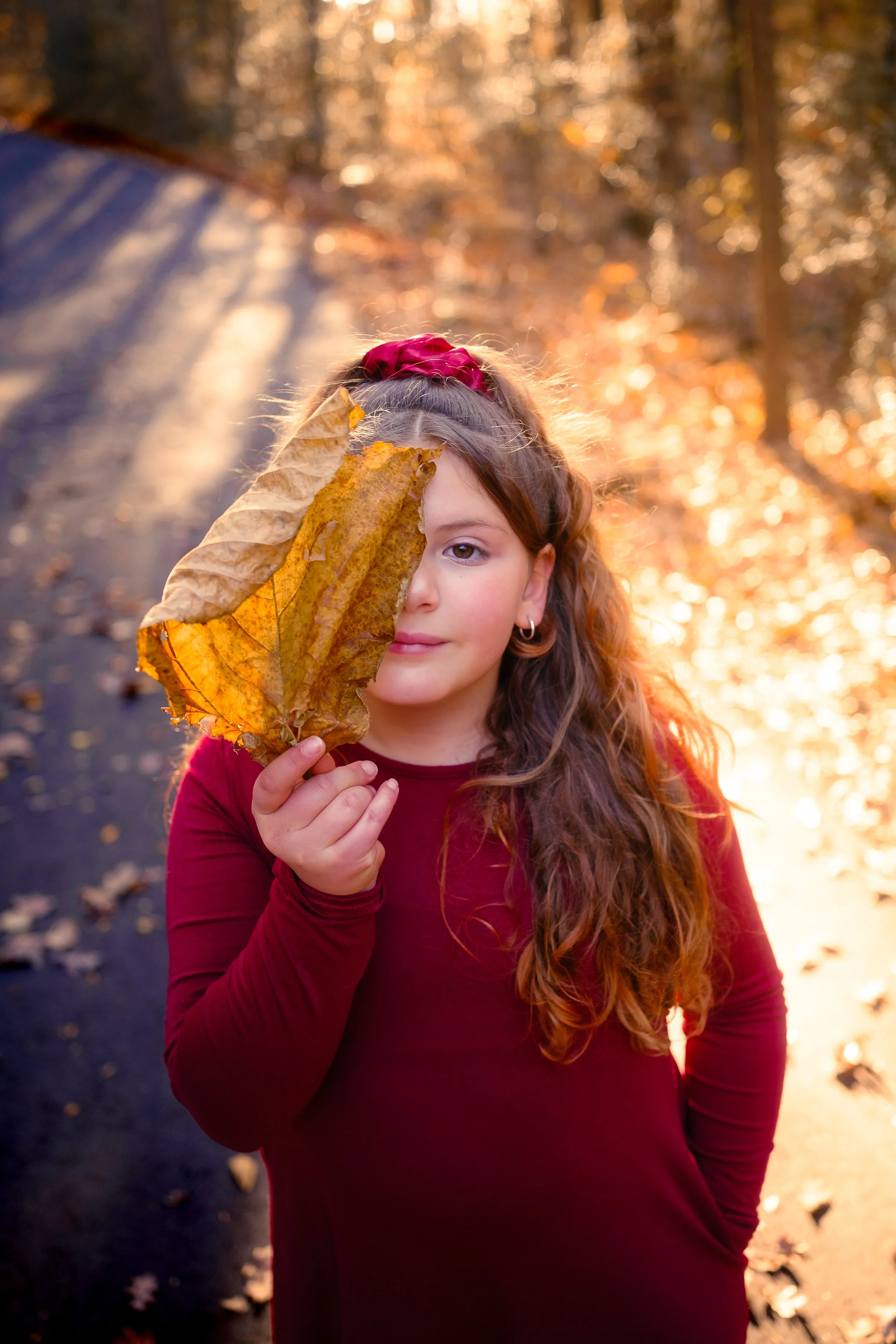 A young girl with long wavy hair, wearing a maroon long-sleeve shirt and a red scrunchie in her hair, holding a large yellow autumn leaf in front of her face, half covering her eye, on a wooded path during fall with golden sunlight filtering through 