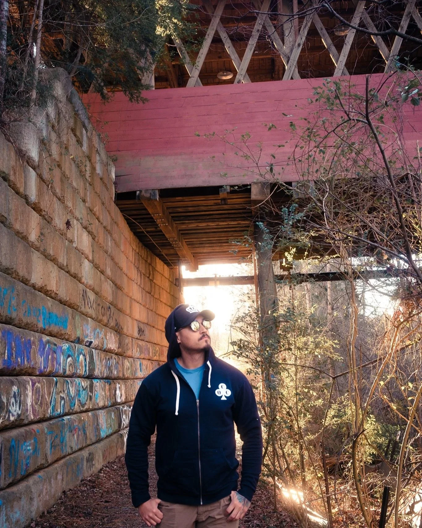 Man in black hoodie and sunglasses standing by graffiti-covered brick wall and wooded area at sunset.