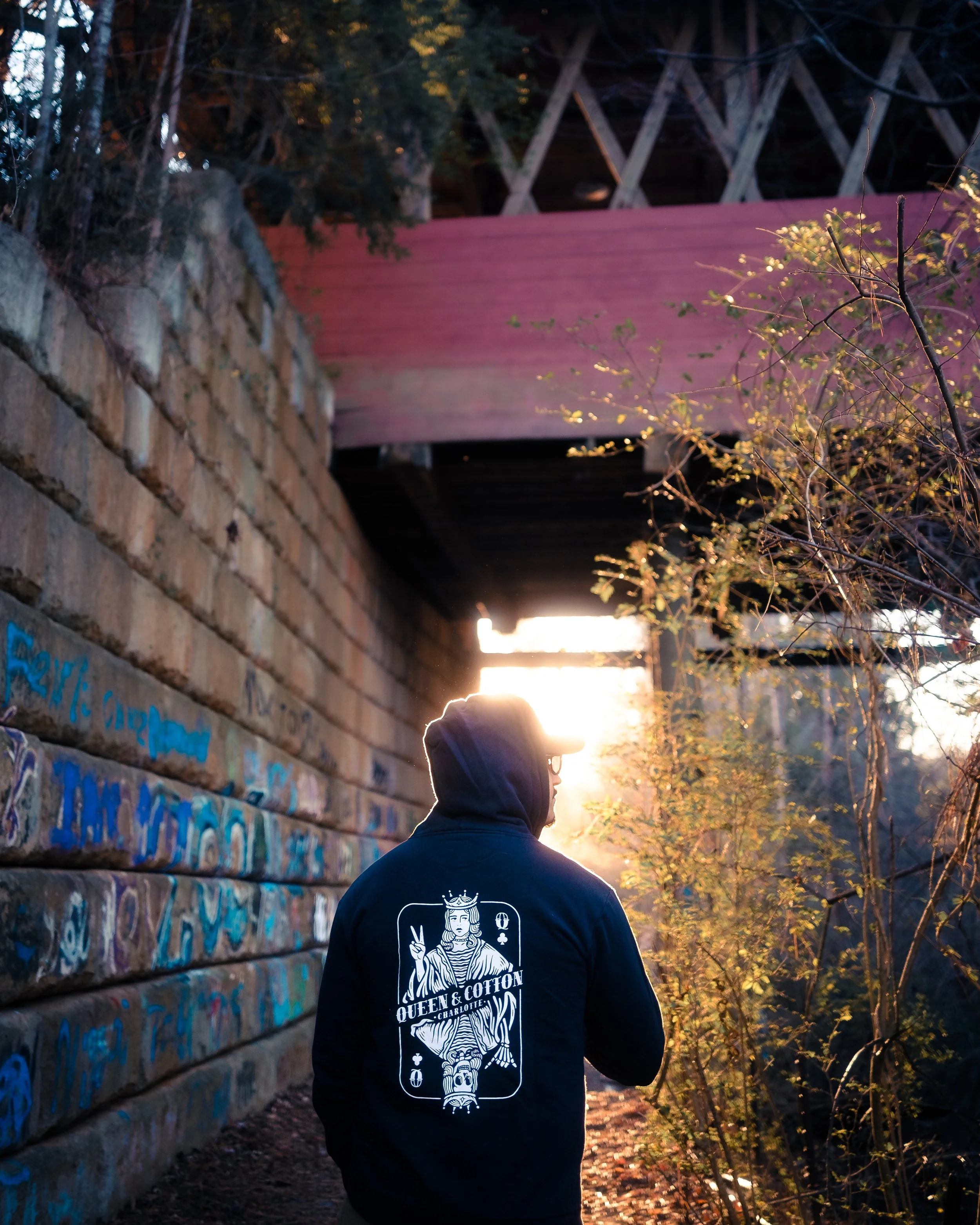 A person wearing a black hoodie with Queens & Cotton text and graphic, standing in a graffiti-covered alleyway with trees and a bridge overhead, during sunset.