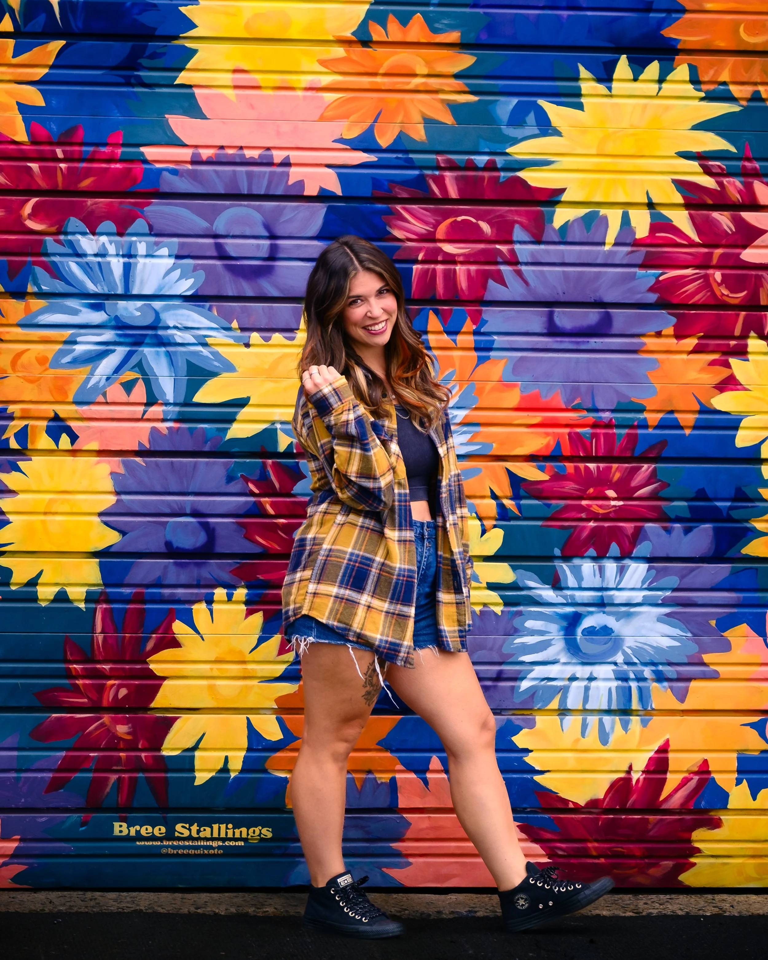 A young woman posing in front of a colorful mural of large, vibrant flowers on a garage door.