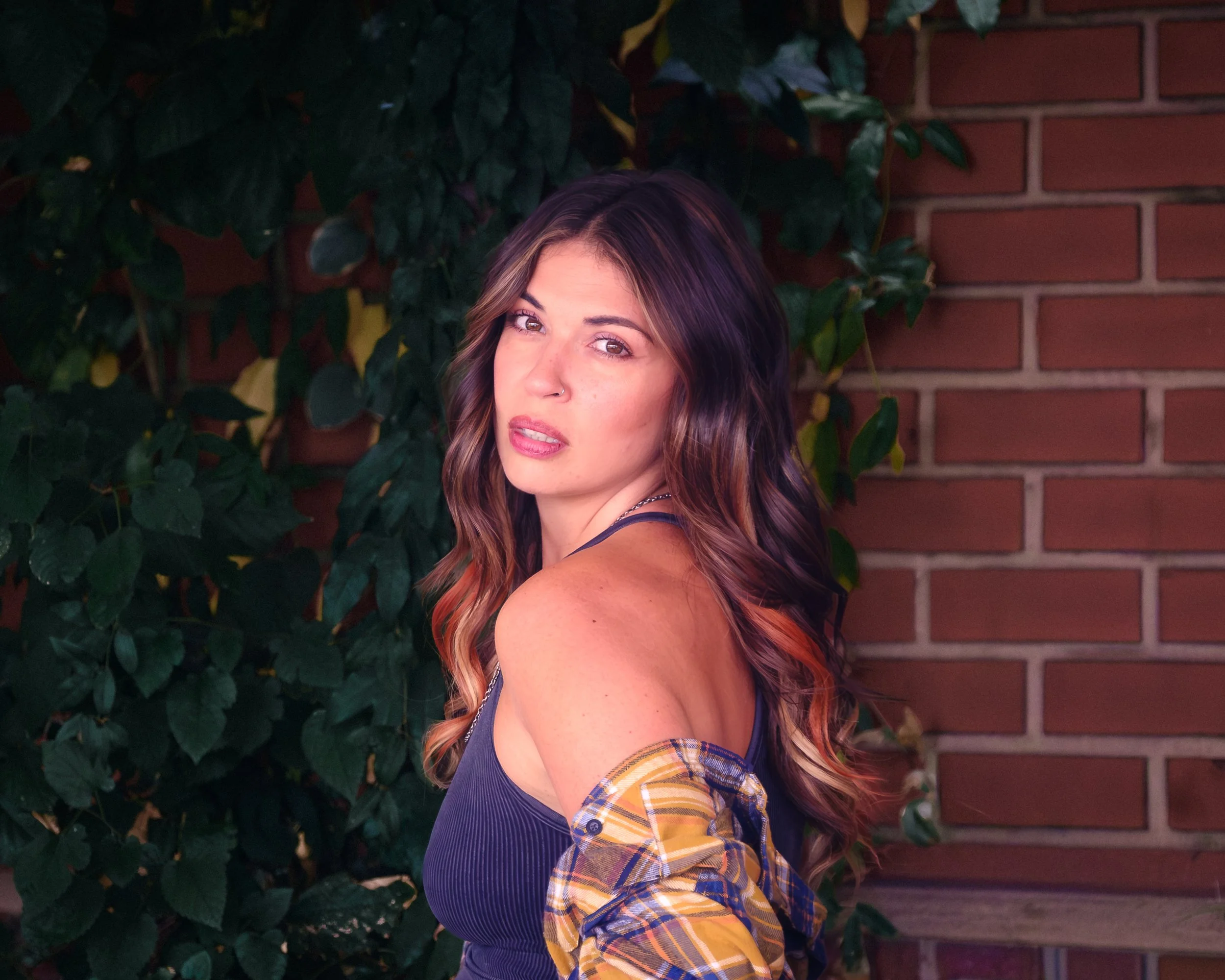 A woman with wavy brown hair with highlights, wearing a black top and a plaid shirt draped over her shoulders, standing outdoors near a brick wall and green foliage, looking at the camera with a serious expression.