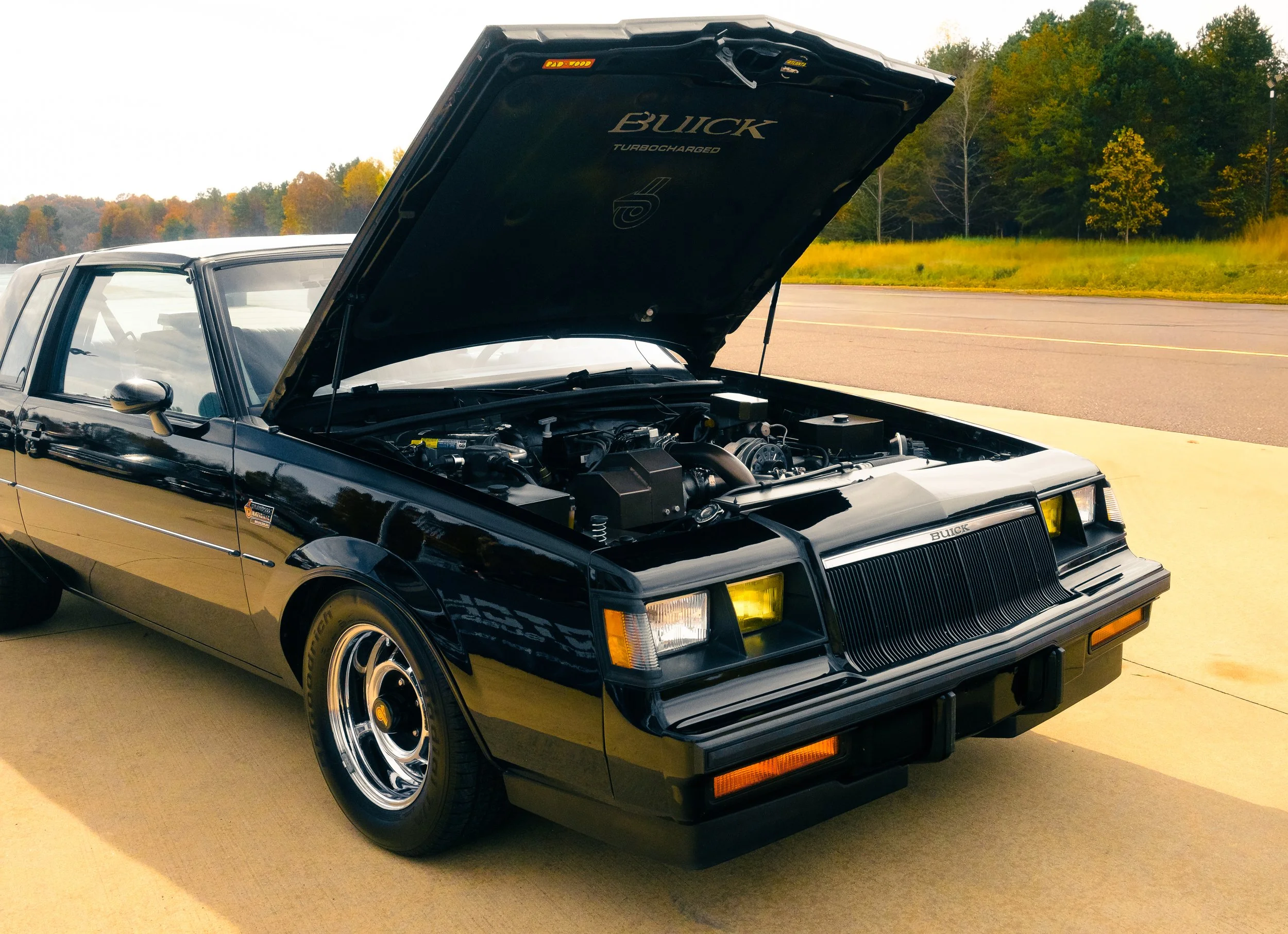A classic black Buick car with the hood open, revealing its engine, parked on a light-colored concrete surface with trees in the background.