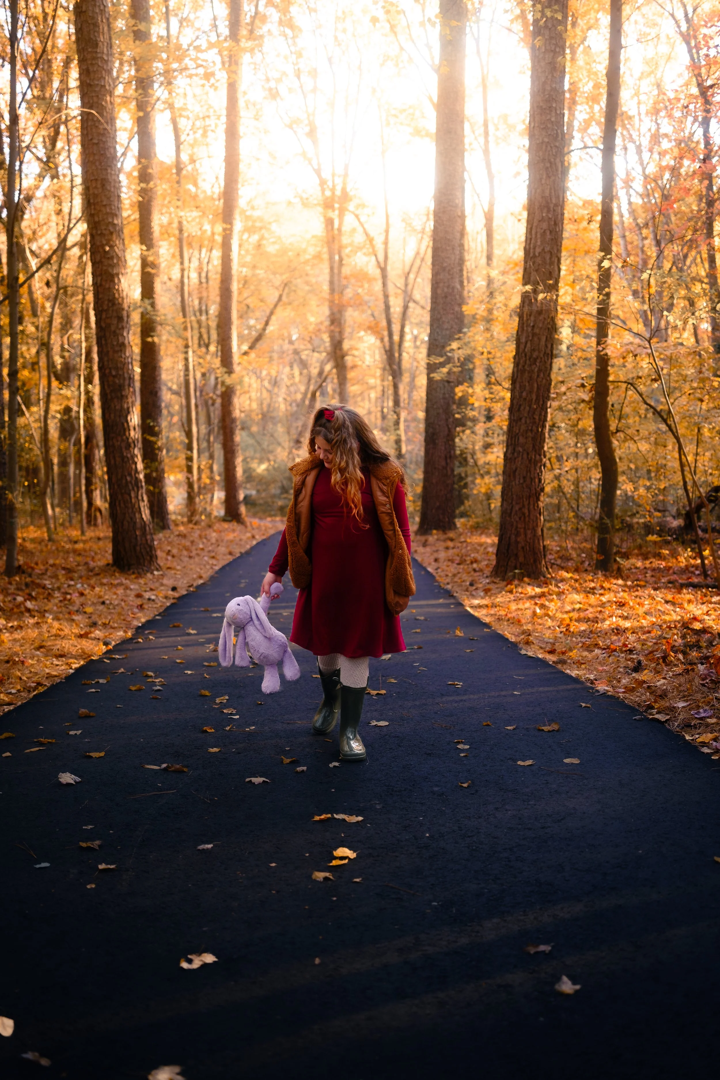 A woman walking on a paved forest trail during autumn, holding a purple stuffed bunny. She is wearing a red dress, a brown vest, and rain boots, with fall foliage and tall trees illuminated by sunlight.