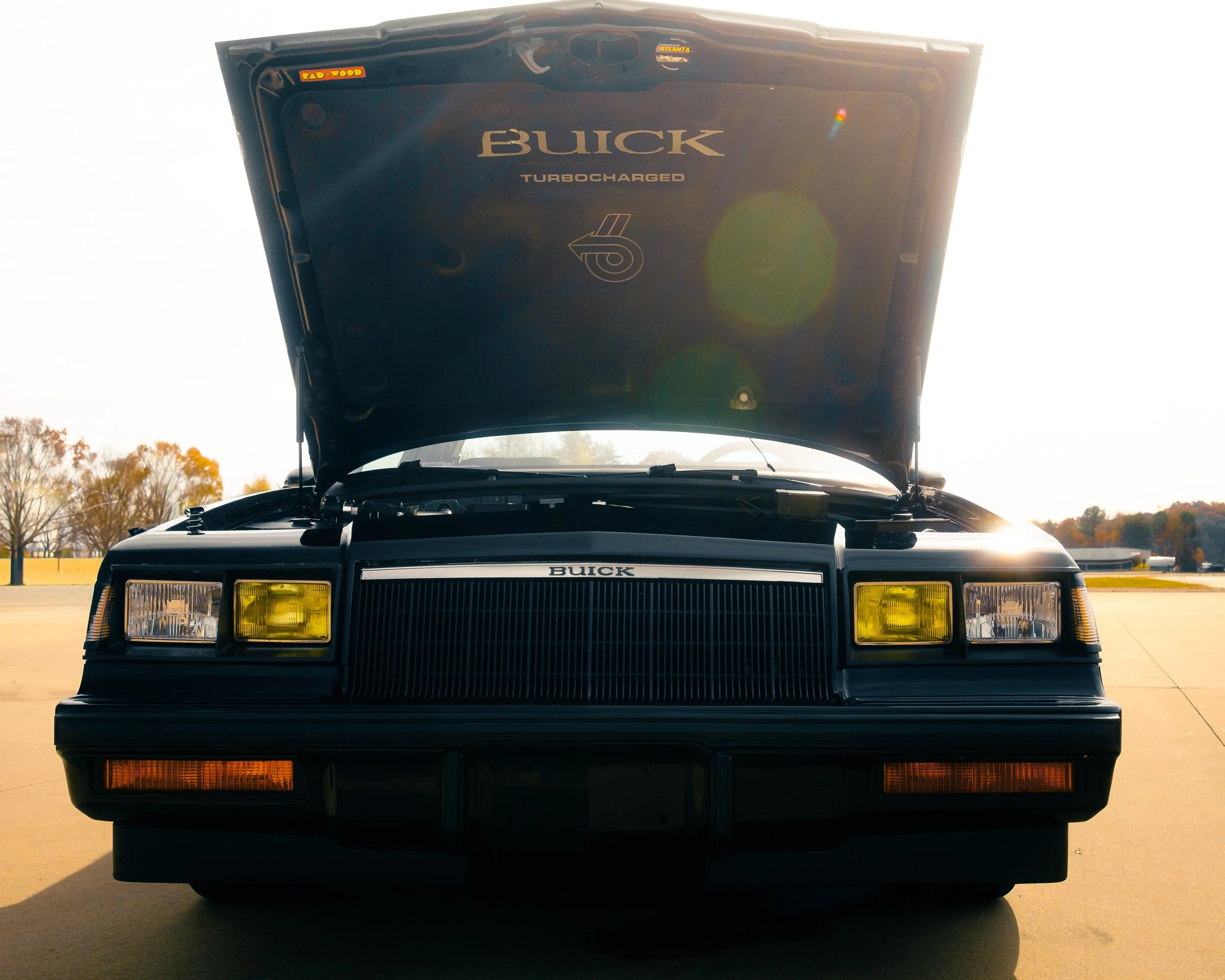 Front view of a black Buick car with its hood open, showcasing the engine compartment, parked outdoors with trees in the background.