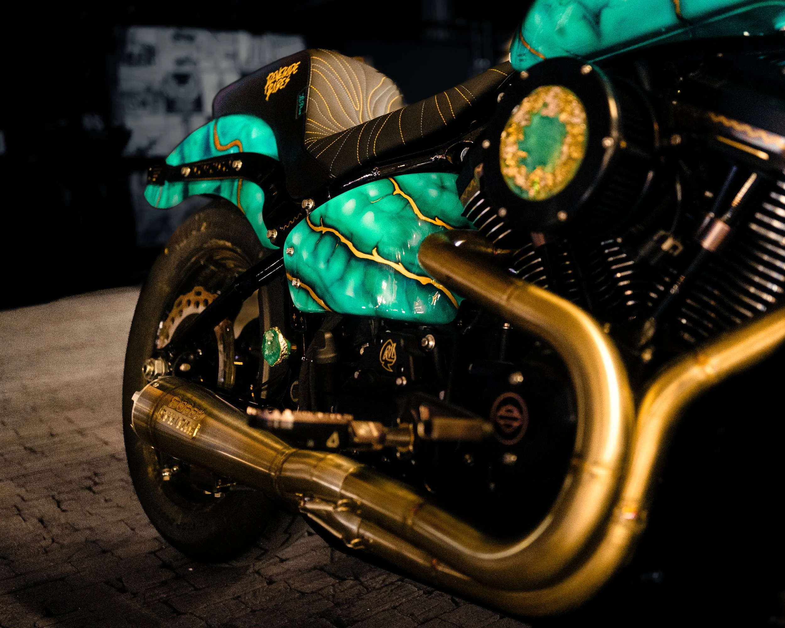 Close-up of a motorcycle with a custom teal and gold painted body, gold exhaust pipes, and a black seat.