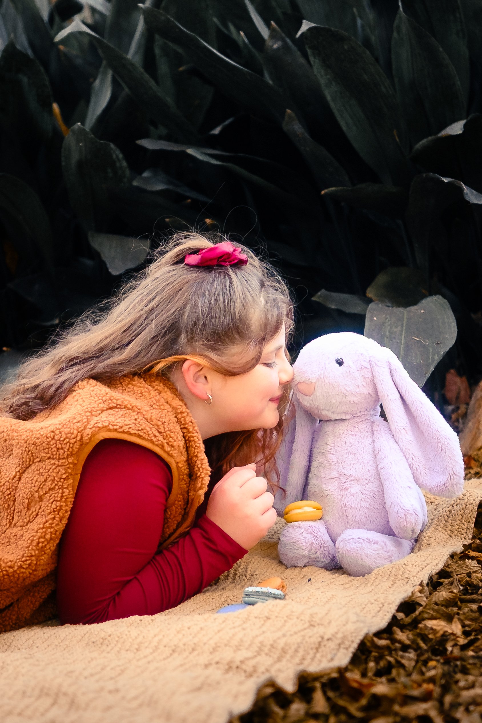 A girl with brown hair and a red scrunchie, wearing a red long sleeve shirt and a brown fleece vest, is lying on her stomach on a beige blanket outdoors. She is face-to-face with a plush purple bunny toy while holding colorful macarons. The backgroun