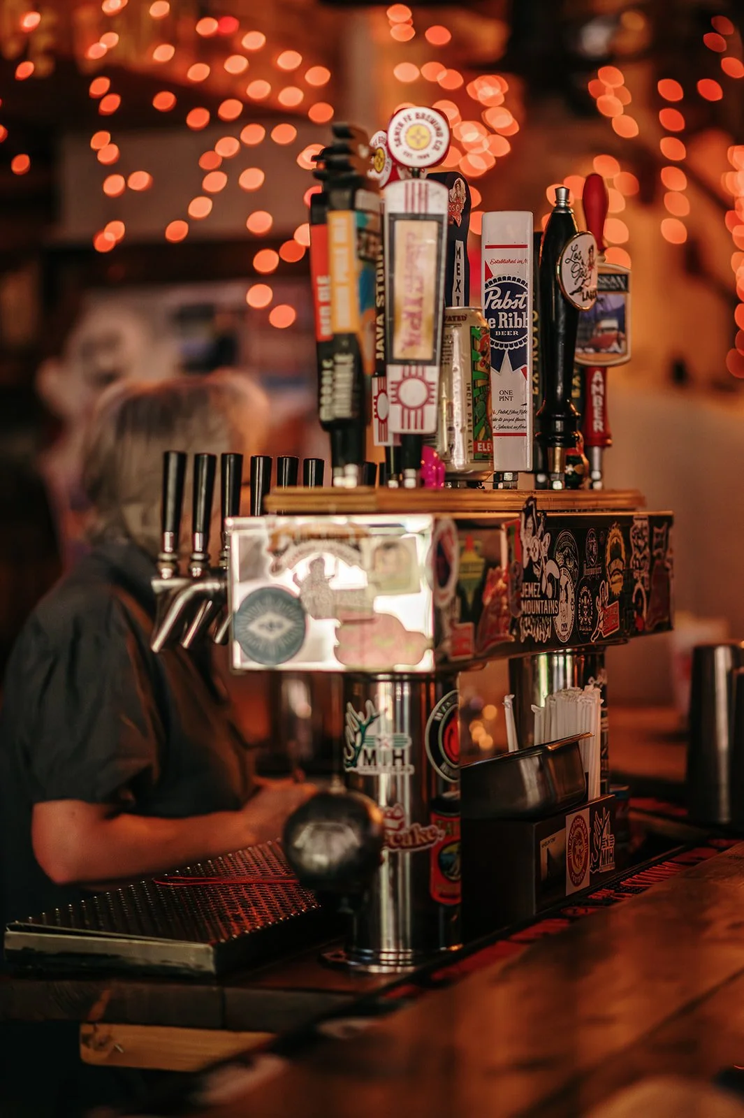 Close-up of a bar beer tap system with various beer taps and a bartender in the background, blurred, in a dimly lit bar with orange string lights.
