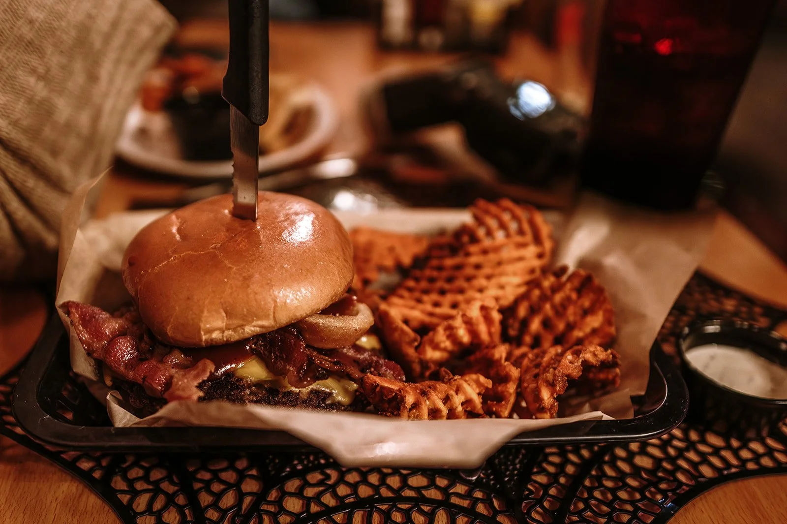 A burger with bacon and lettuce on a bun, served with waffle fries and dipping sauce on a black tray.