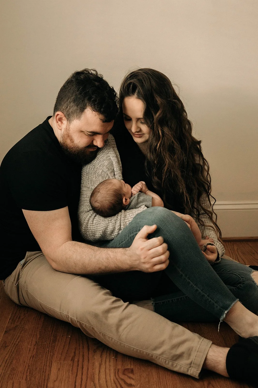 A couple sitting on a wooden floor with their newborn baby, cradling the baby and looking at it lovingly.