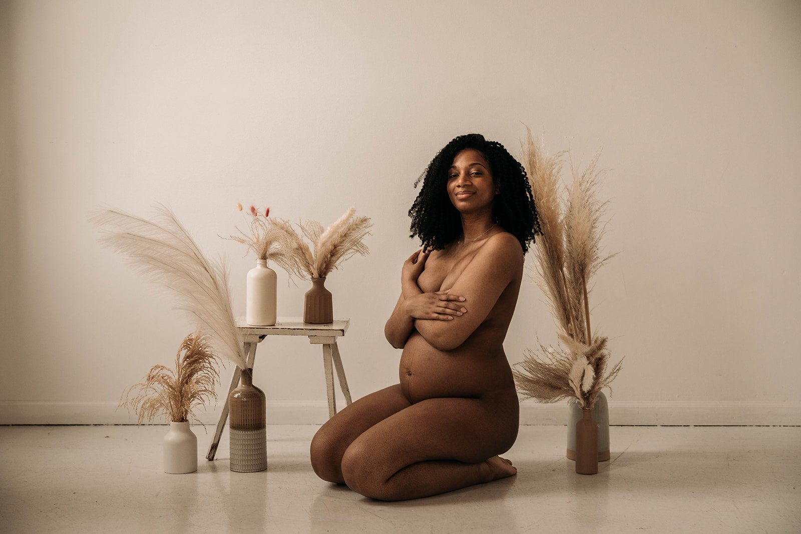 A nude woman kneeling on the floor with arms crossed over her chest, in front of a minimalist backdrop with vases of dried pampas grass.