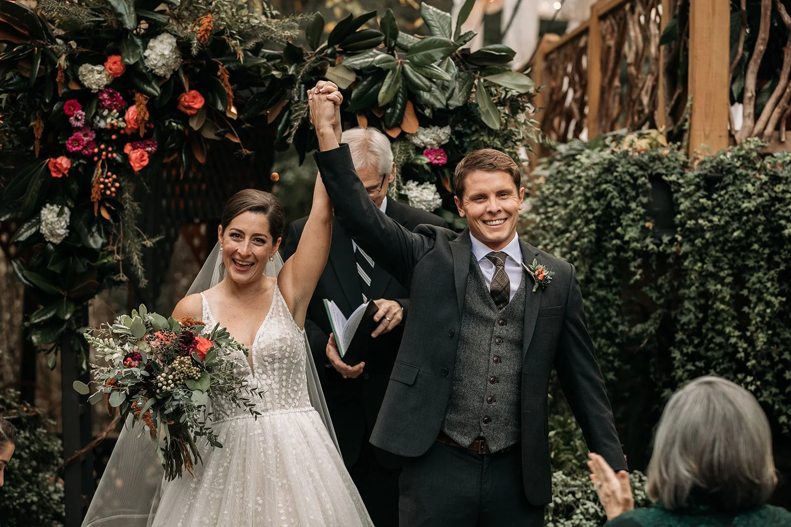 A newly married couple celebrating their wedding ceremony outdoors, with the bride wearing a white dress and holding a bouquet of flowers, and the groom in a dark suit. They are smiling and holding hands up in victory as they are being watched by gue