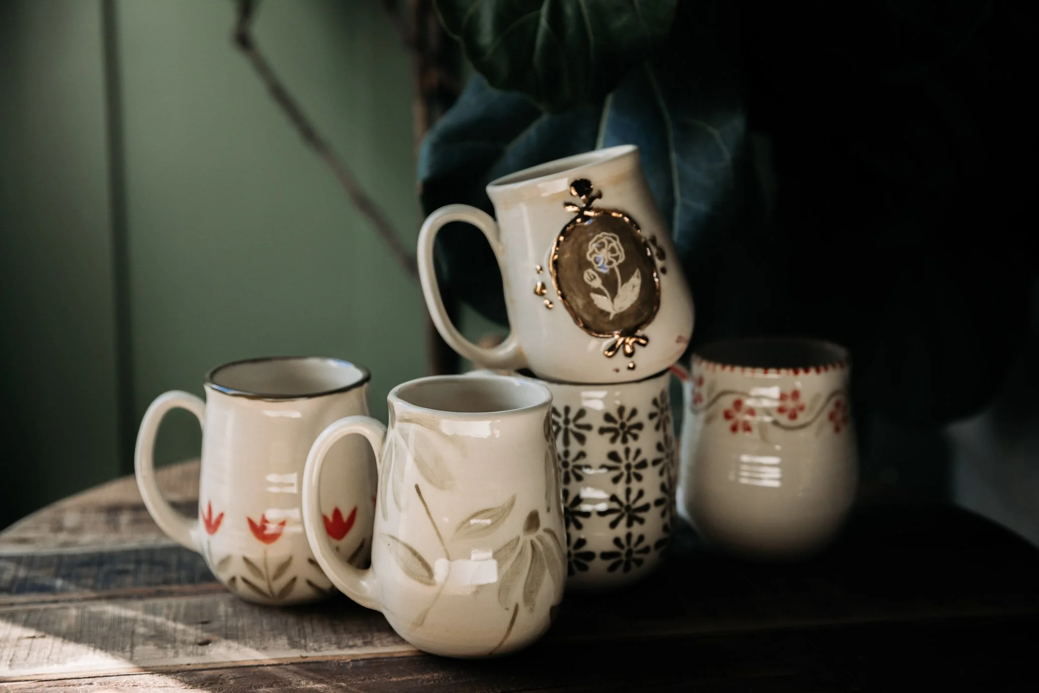 Collection of five ceramic mugs with floral and decorative patterns arranged on a wooden surface, with a green wall background and some dark foliage.