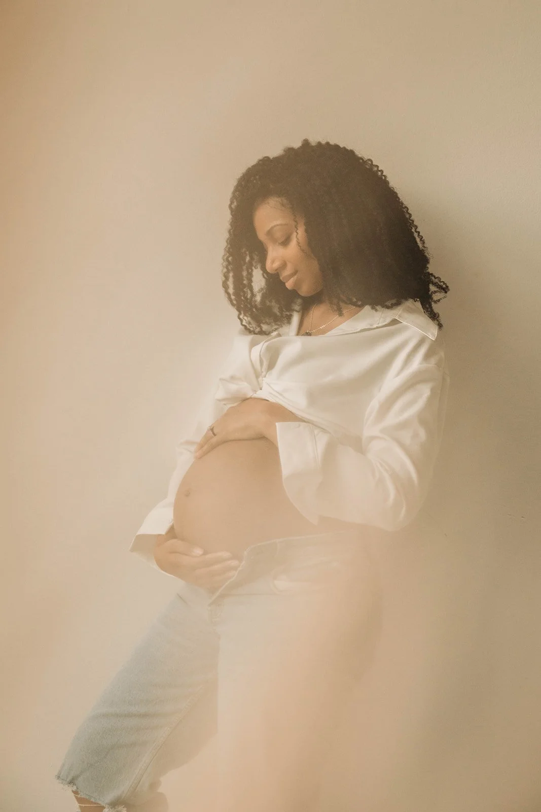 A pregnant woman with curly hair wearing a white satin shirt and jeans, gently holding her belly and standing against a neutral background.