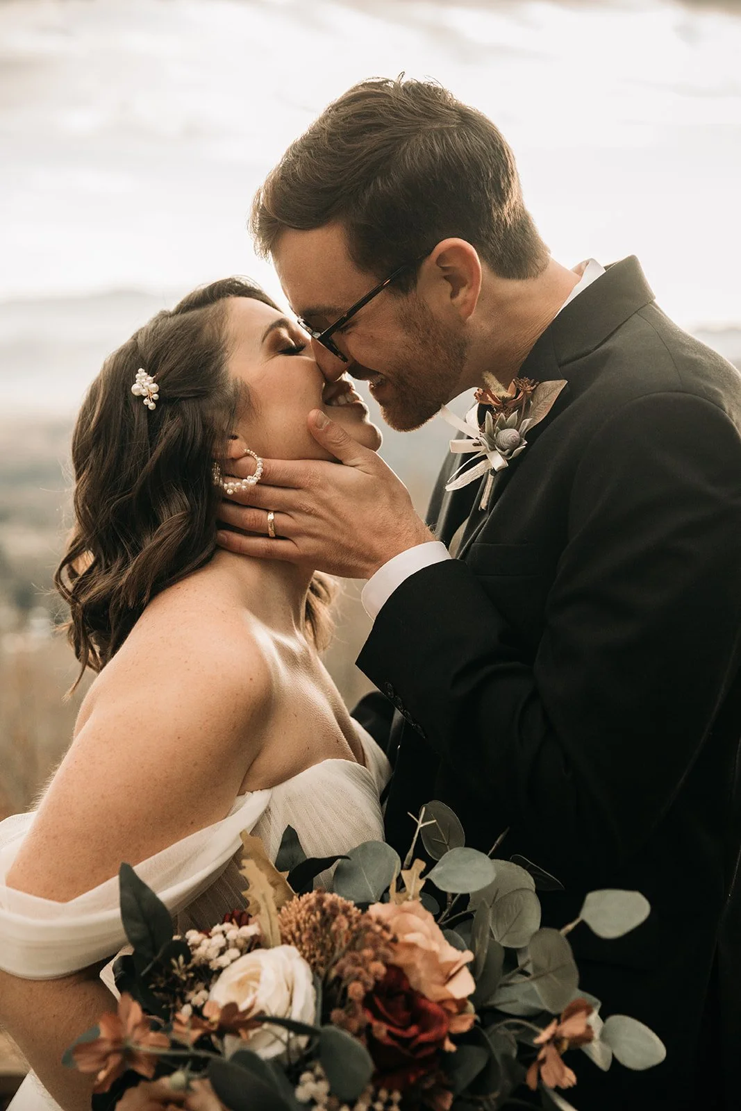 A bride and groom share a kiss on their wedding day at the beach, with the groom holding the bride's face and the bride holding a bouquet of flowers.