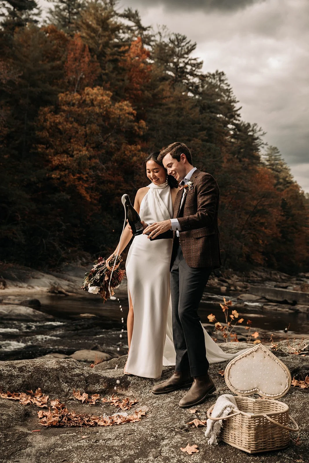 A bride and groom celebrating outdoors on a rocky riverbank surrounded by autumn-colored trees. The bride is in a white dress, holding a bouquet, and the groom is in a brown blazer and dark pants. They are smiling while opening a bottle of champagne with spilled liquid around them. A wicker basket with a cloth is on the ground nearby.