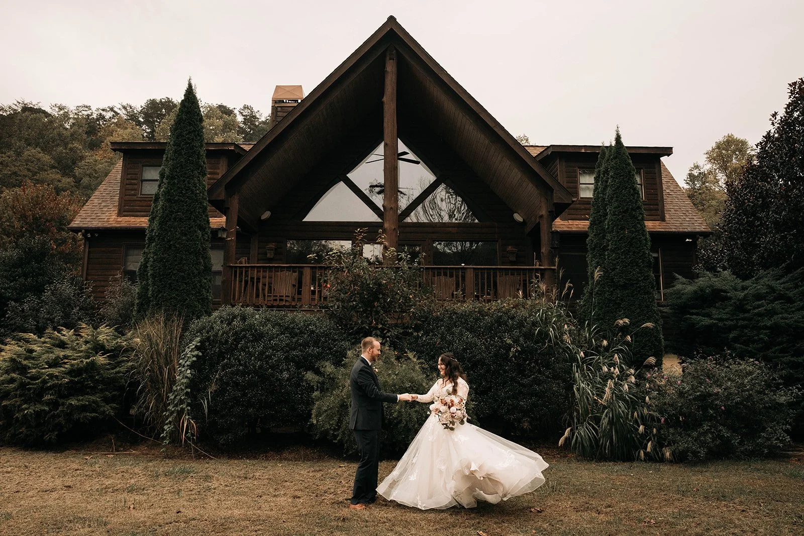 A bride and groom holding hands in a garden outside a large wooden house, with the bride in a white wedding gown holding a bouquet, and the groom in a dark suit, all surrounded by tall trees and bushes.
