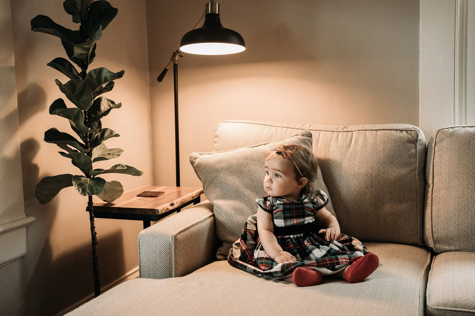 A young girl sitting on a beige sofa, wearing a plaid dress, with a headband, and red shoes. The sofa is in a warmly lit living room with a wooden side table, a leafy houseplant, and a black floor lamp.