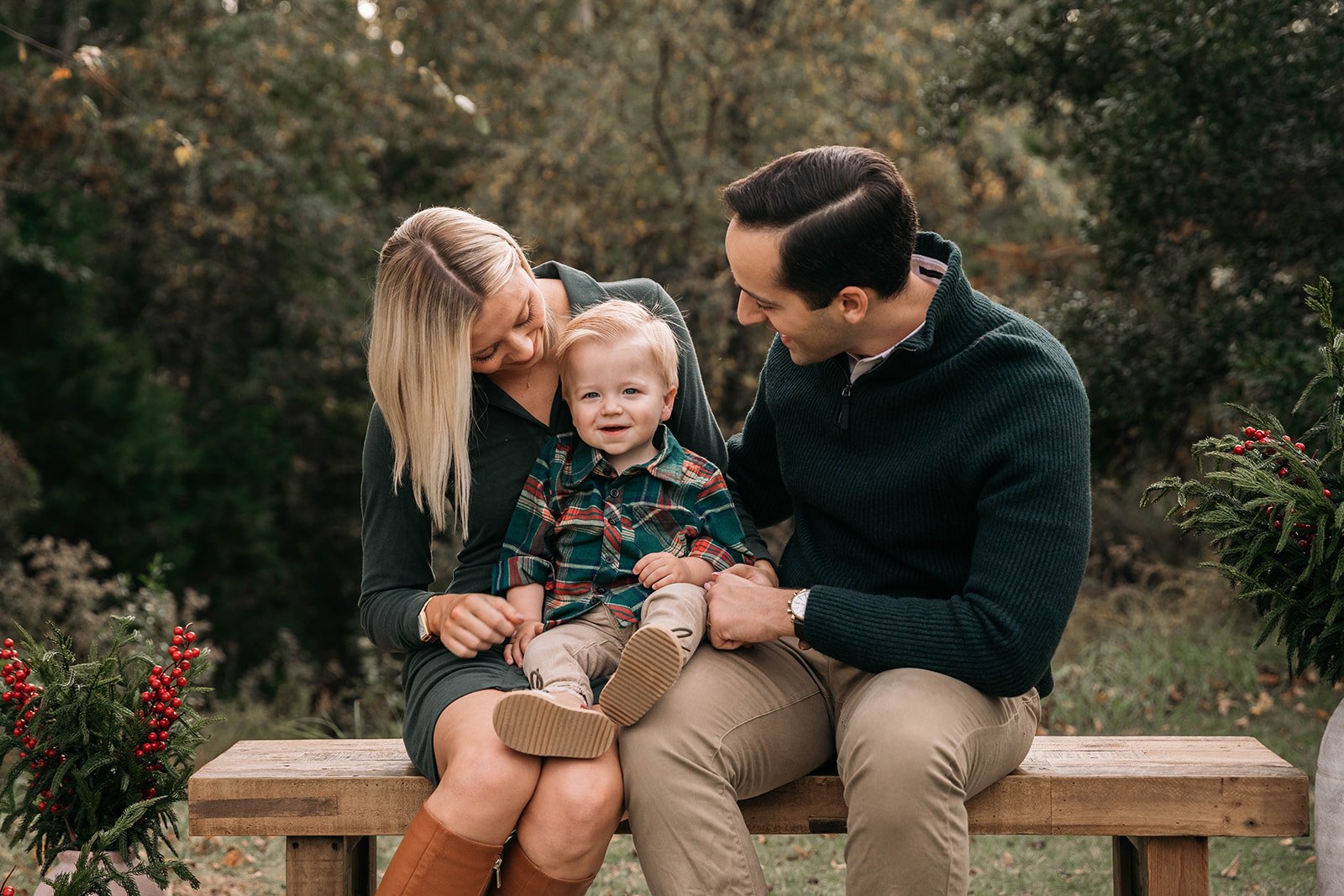 A family sitting on a wooden bench outdoors during fall, with trees and holiday decorations around, featuring a woman, a man, and a young boy smiling and interacting.