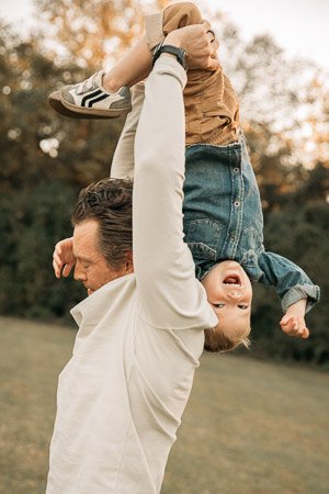 A man lifting a young boy upside down outdoors during fall, with trees and blurred background.