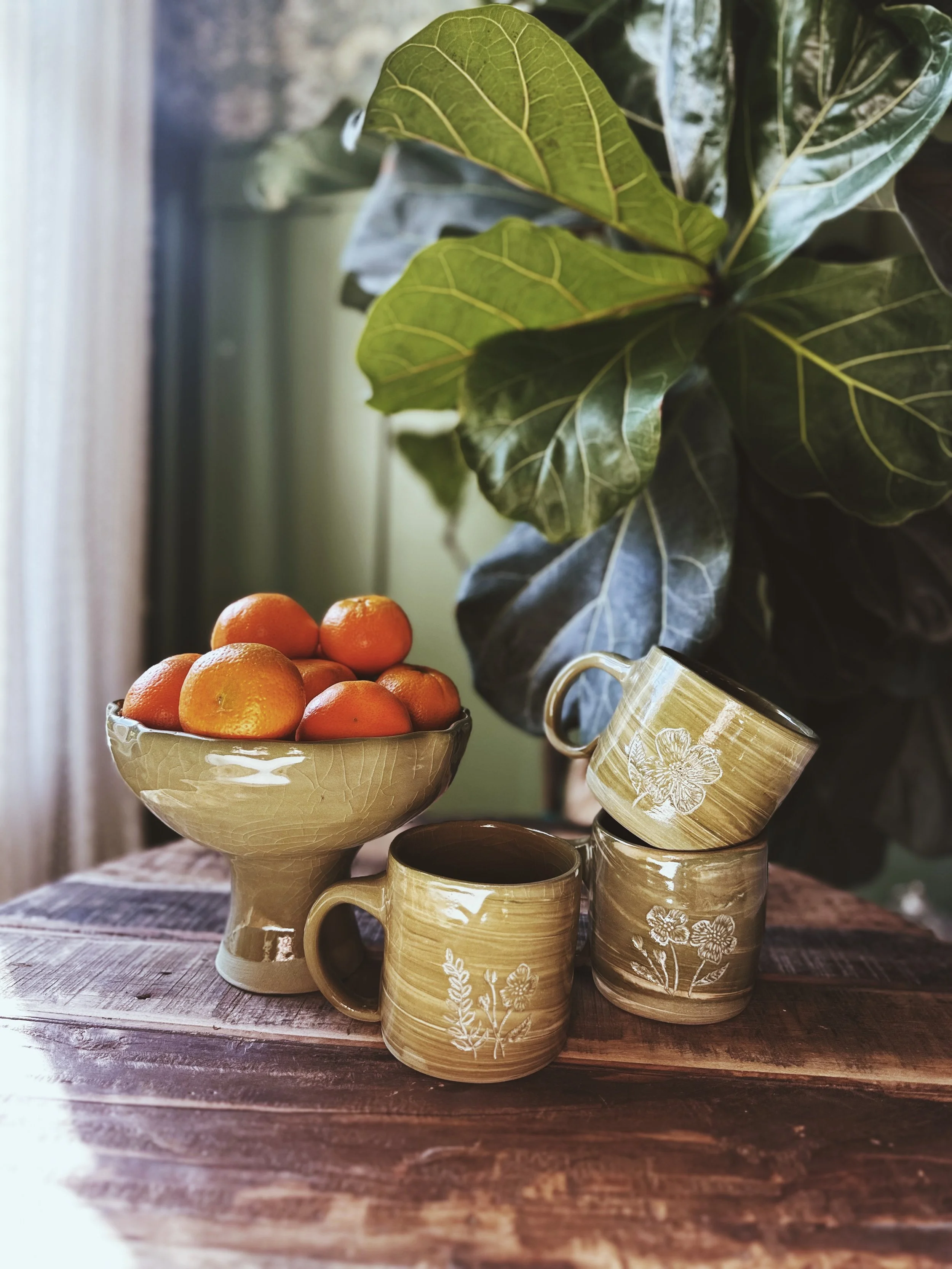 A bowl of oranges and three ceramic cups with floral designs on a wooden surface, with a large leafy plant in the background.